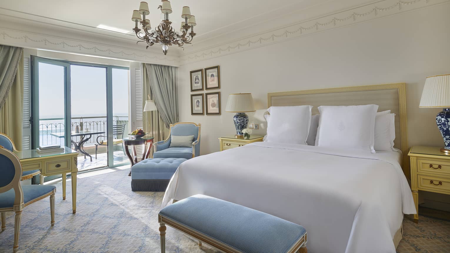 Bedroom with white king bed, chandelier, light blue bench and arm chair, and a balcony overlooking the sea