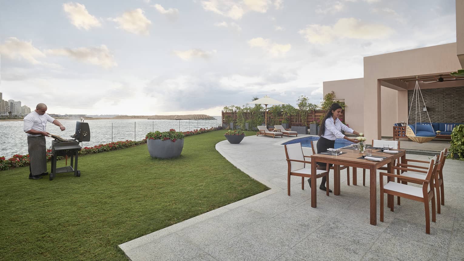 A chef seasons meat on a barbecue near the water’s edge while a server arranges flowers at a table on a patio near a pool.