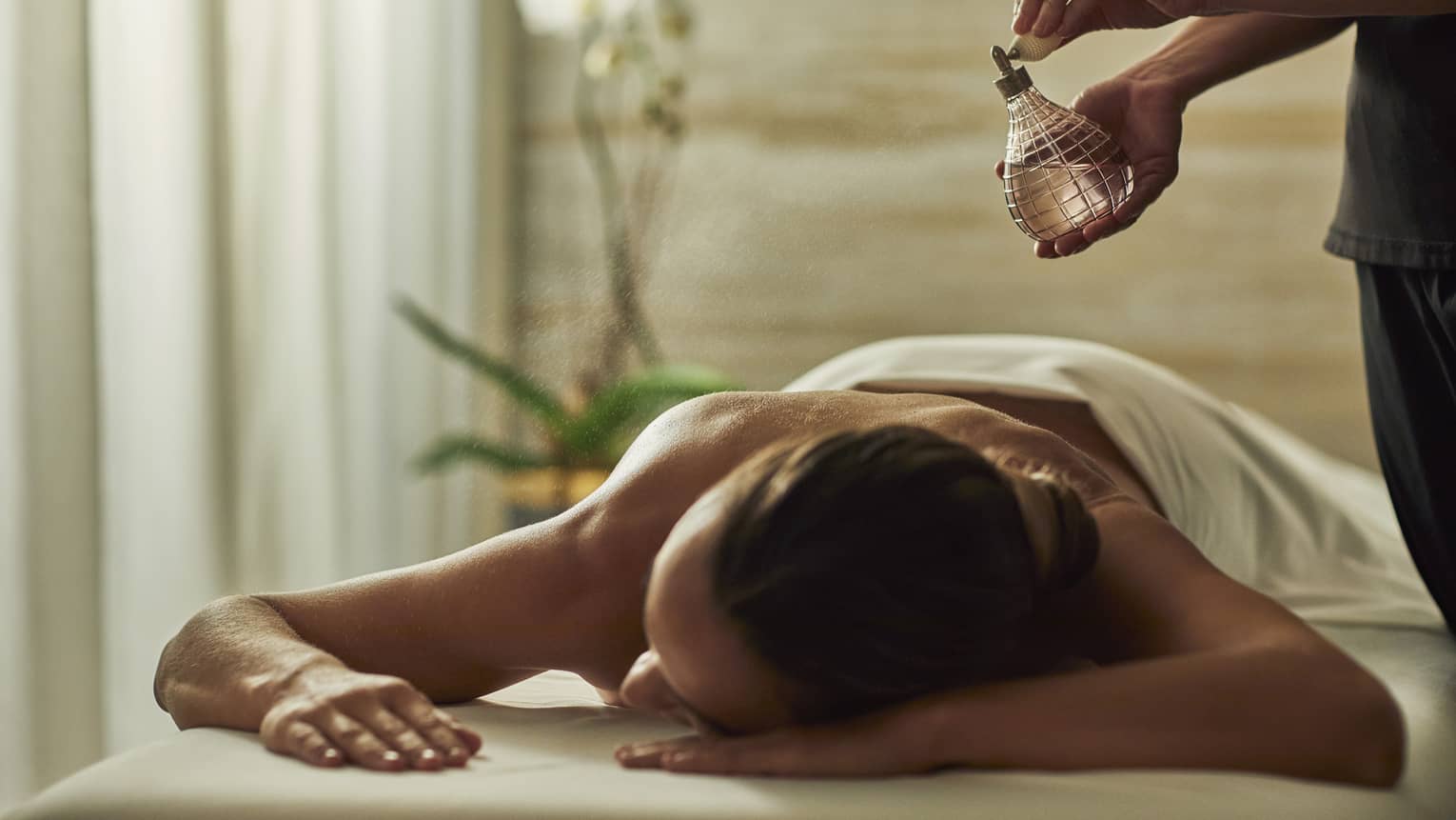 Spa staff sprays perfume bottle over woman lying on massage table