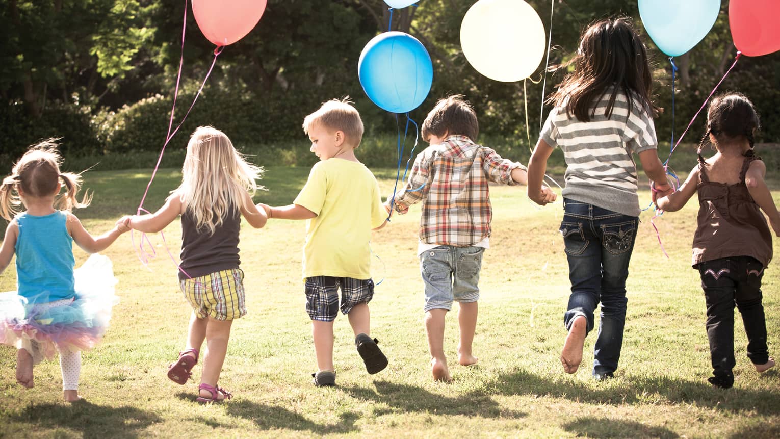 Children walking outside in the sunshine, holding hands, carrying coloured balloons.