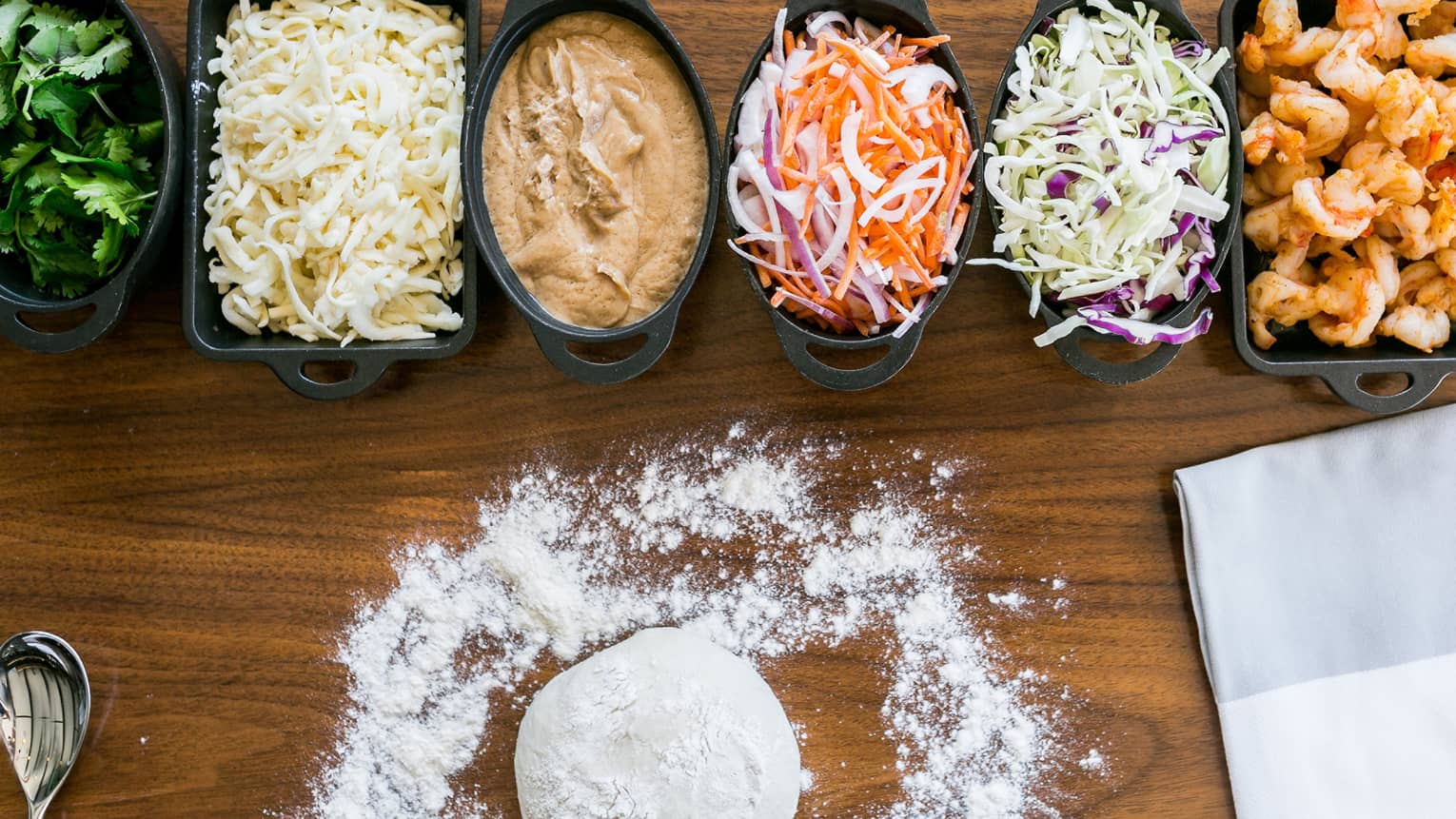Dough and dusted flour on wood counter by row of shredded pizza toppings and sauces in small dishes
