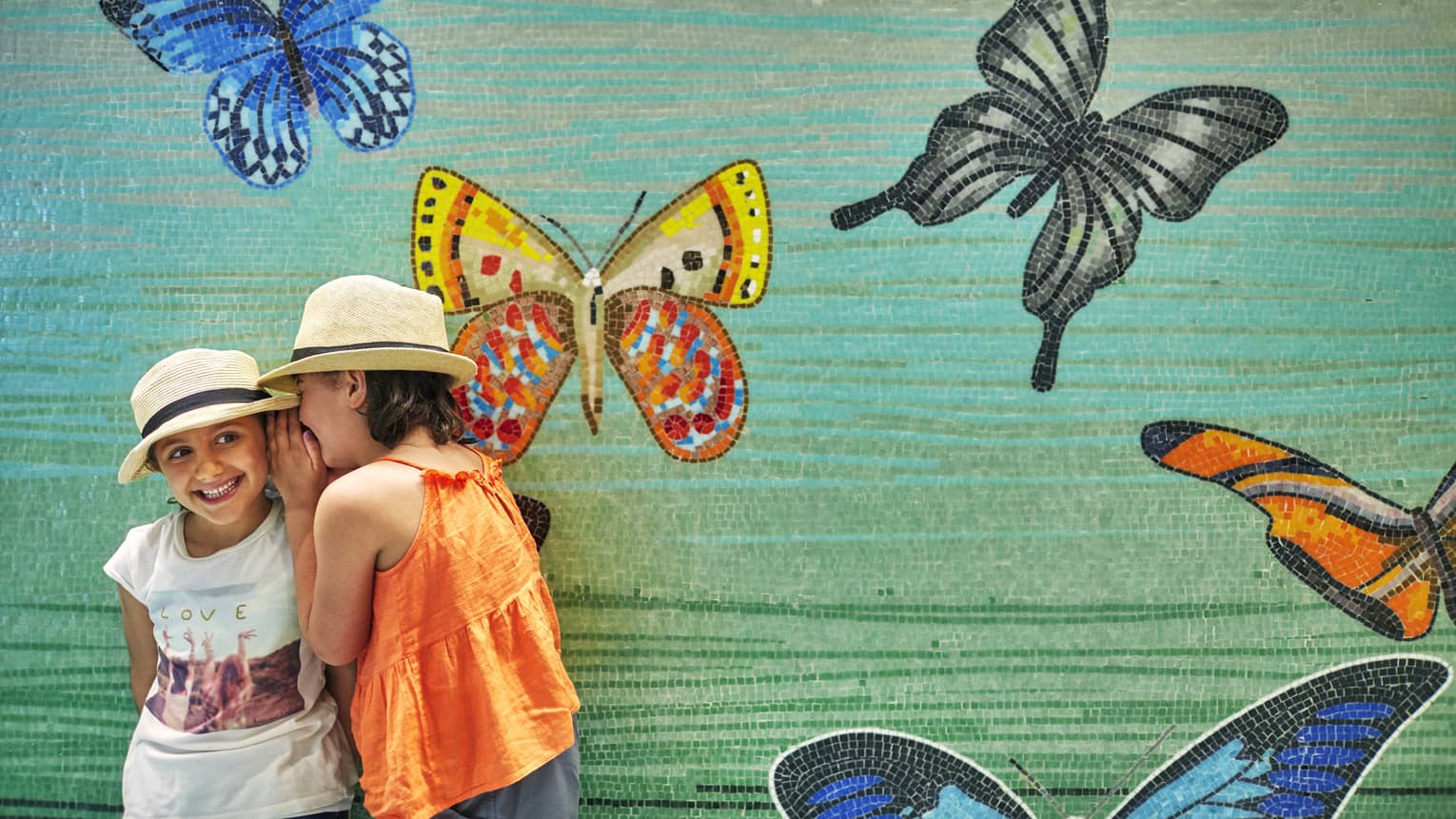 Two young girls whisper in front of butterfly mural wall