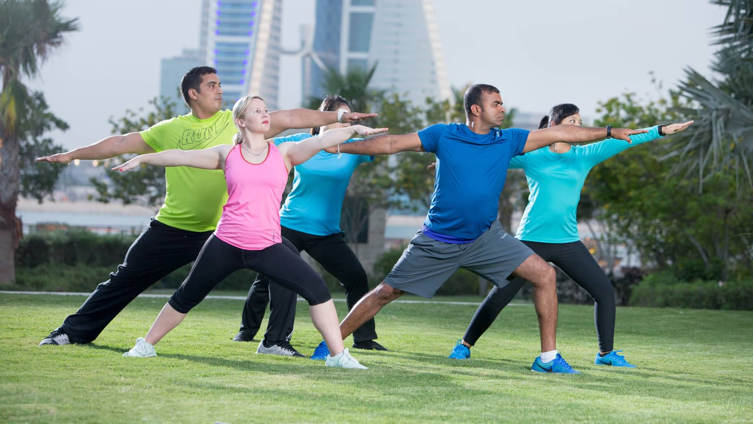 Fitness group stretches in yoga poses on green lawn by water, city skyline