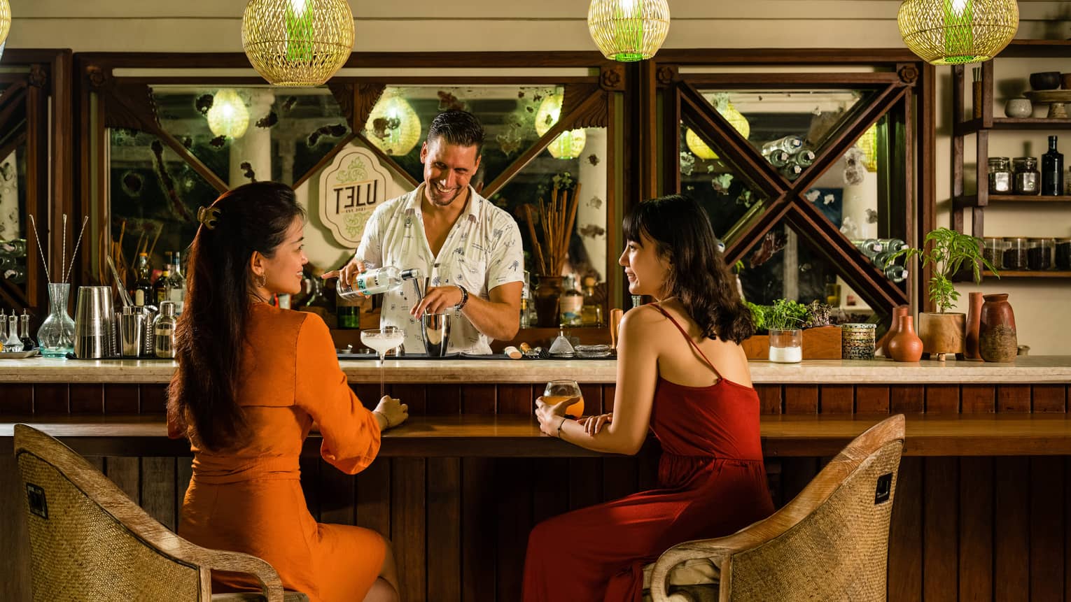 Two women enjoy cocktails at TELU?s bar, smiling male bartender pouring a drink in the background