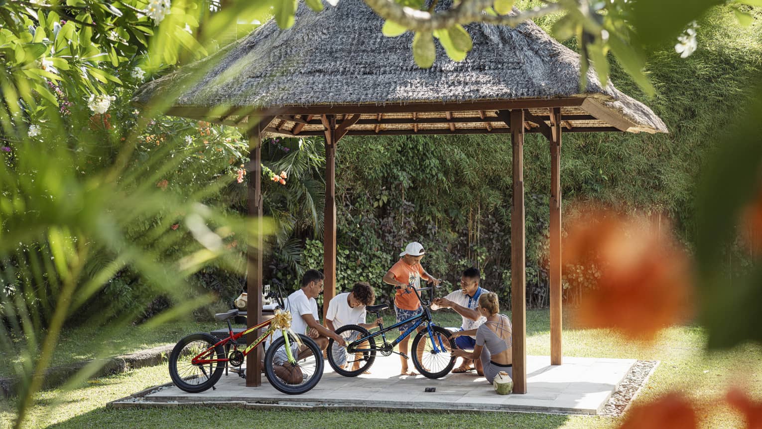 View through foliage of four adults and a child working on a bicycle under a thatched hut surrounded by lush greenery.
