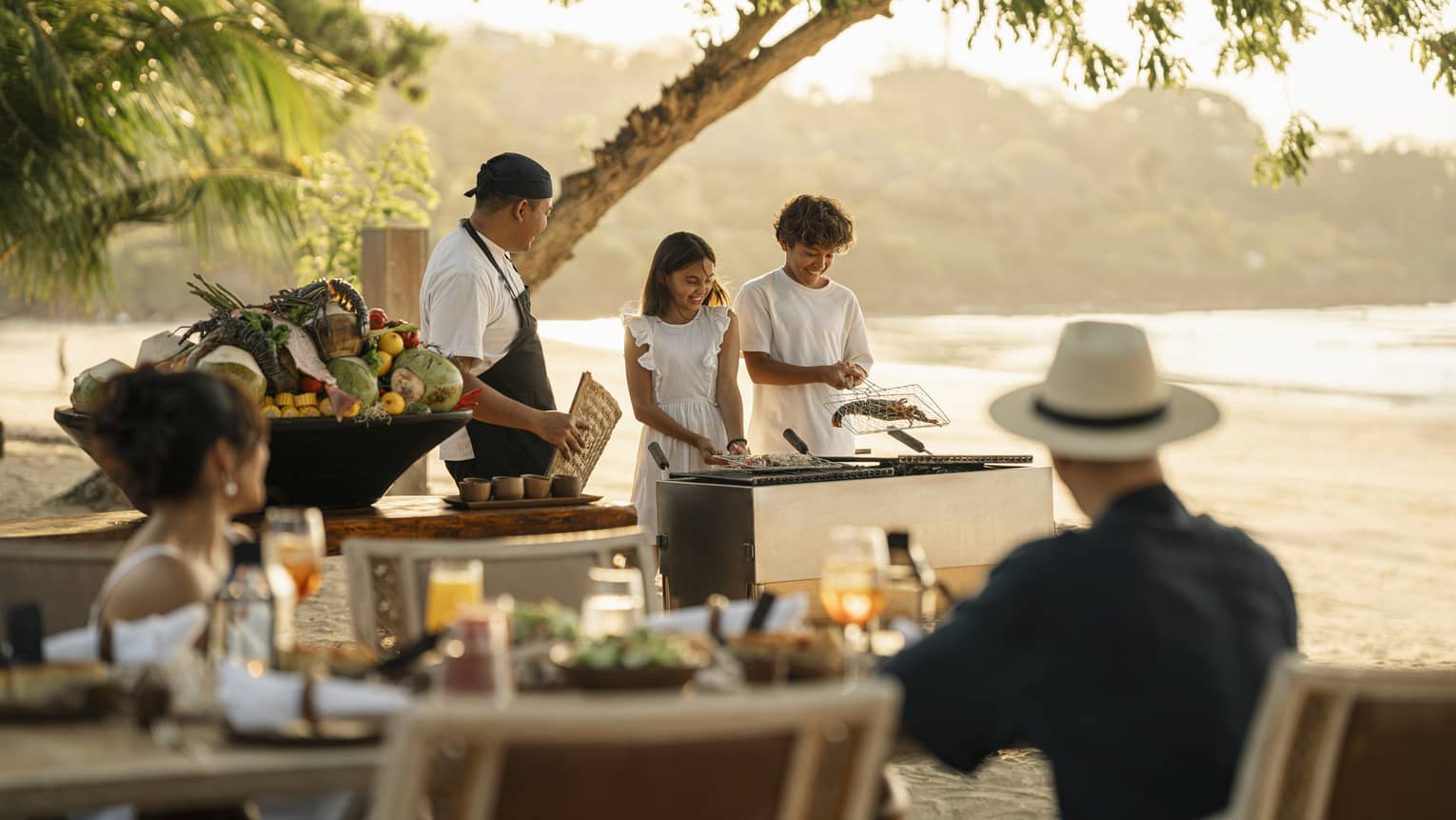 A chef smiles at two children who barbecue shellfish in a grilling basket on the beach as their parents watch.
