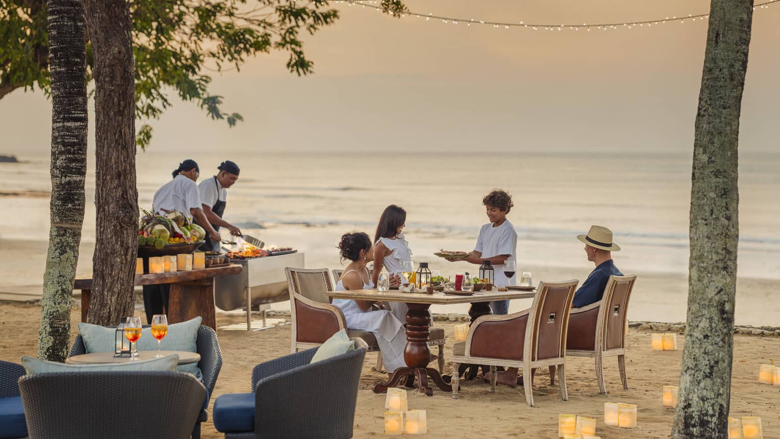 As two chefs barbecue nearby, a child brings a dish to his family’s table on a beach dotted with paper lanterns at dusk.