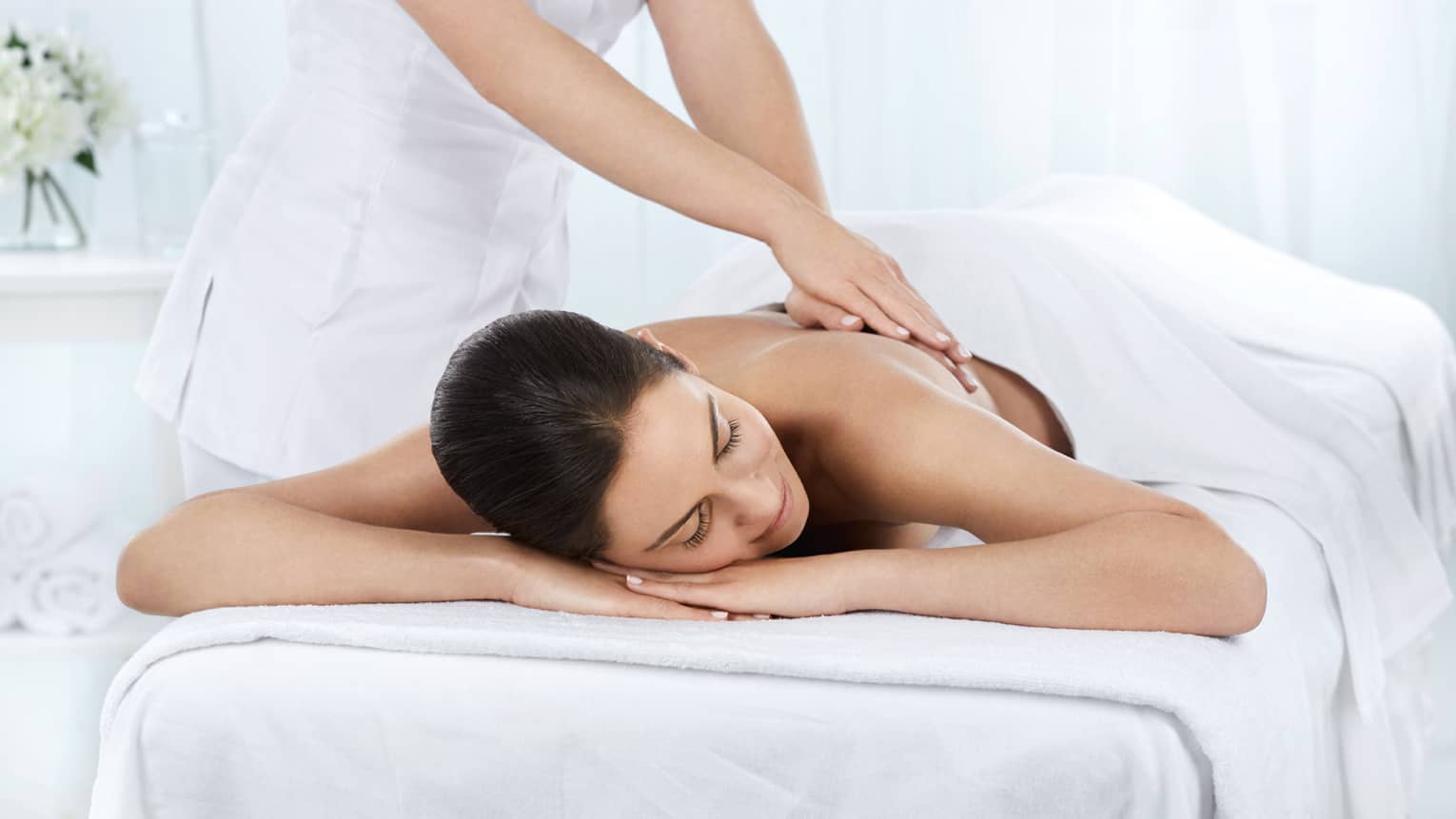 Woman lies under white sheet on massage table as hands massage her bare back