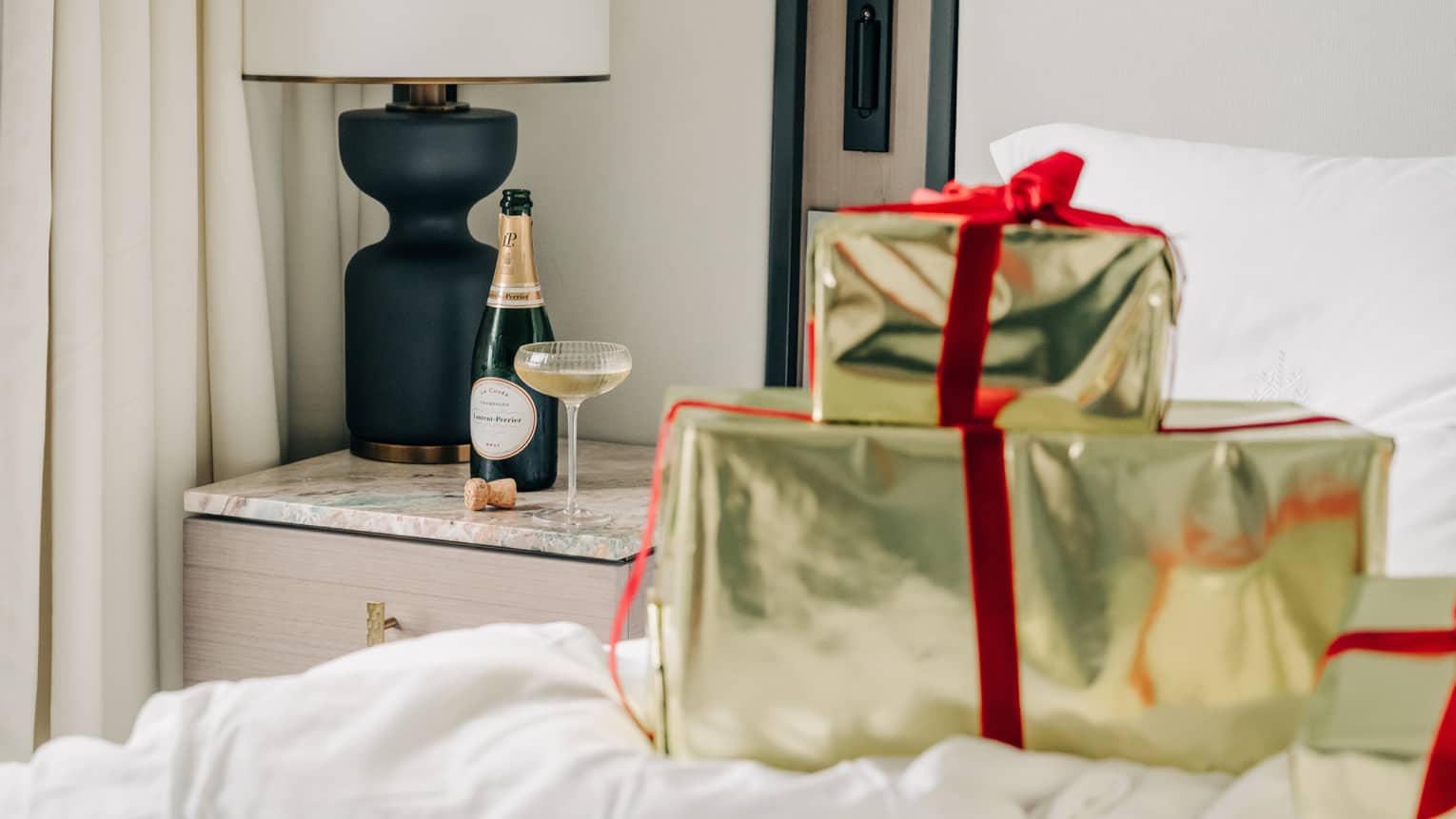 Two shiny gold packages tied up with red velvet ribbon sit on top of a hotel bed with a bottle of champagne and a coup glass set on the nightstand