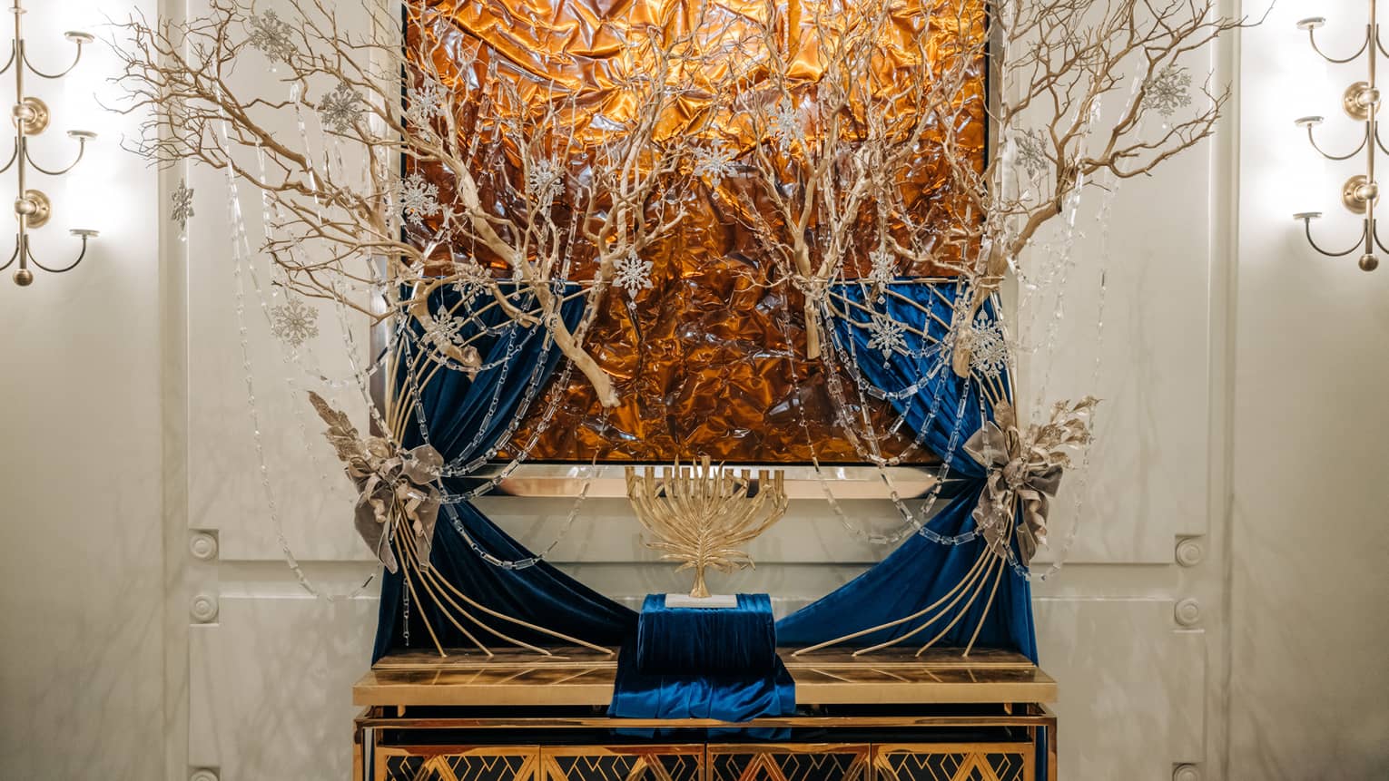 Golden menorah displayed on a blue velvet stand in front of golden tree branches draped in strings of silver