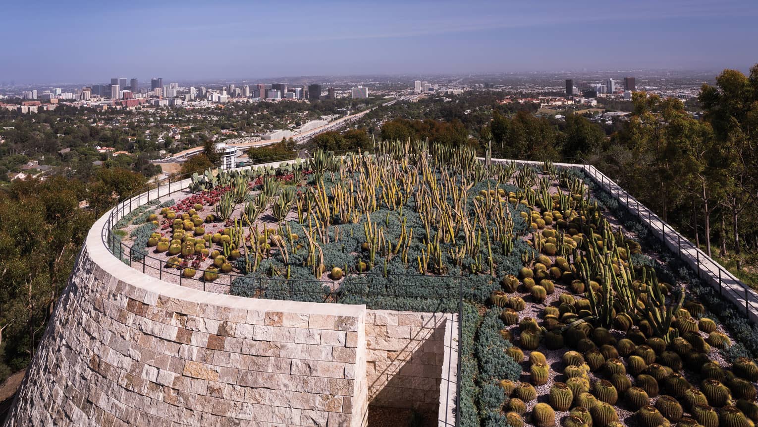 A cylindrical stone structure, topped with a cacti and desert plant garden, towers in the foreground of an urban landscape.