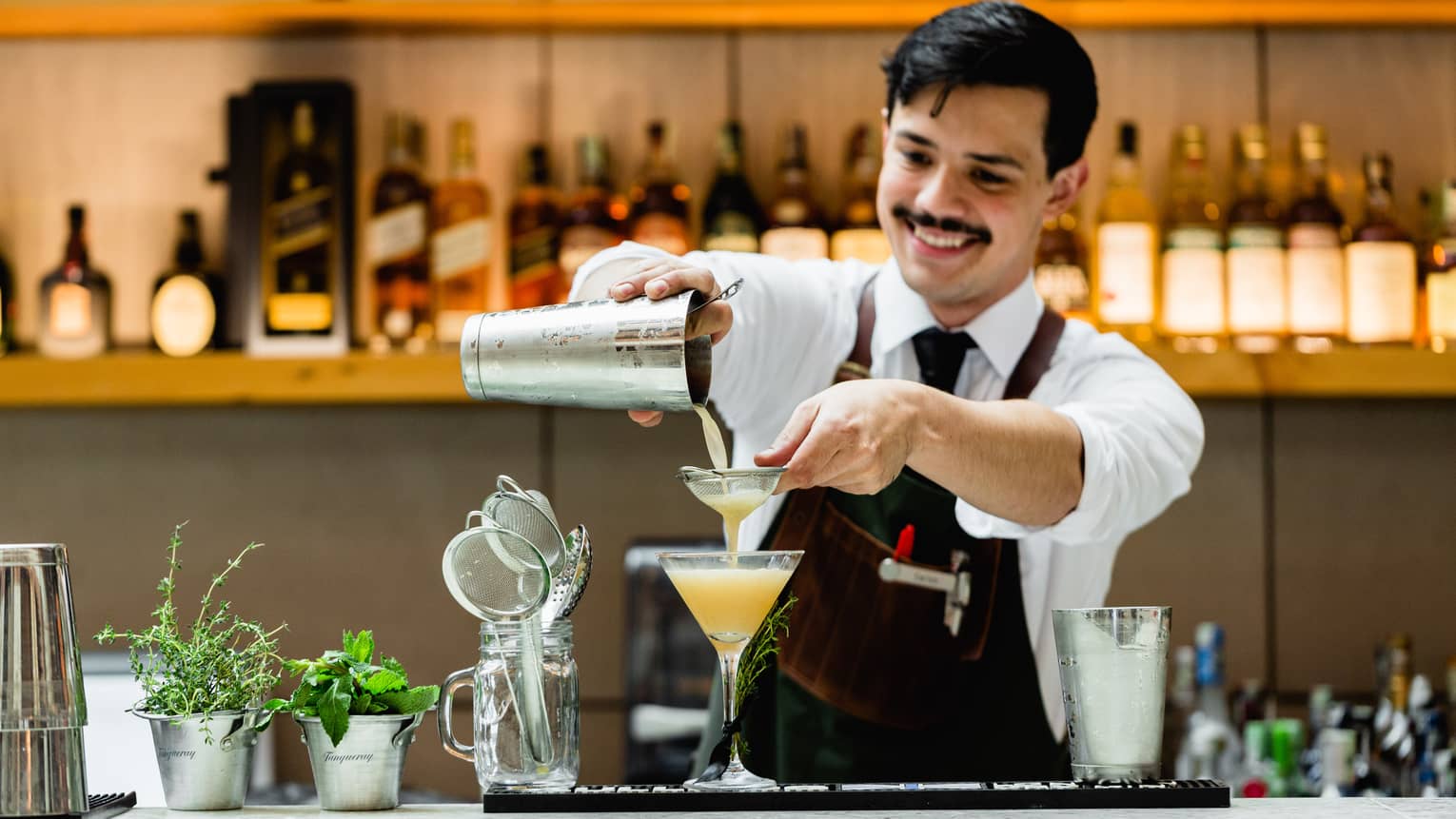 Smiling bartender pours orange cocktail from shaker into martini glass at bar 