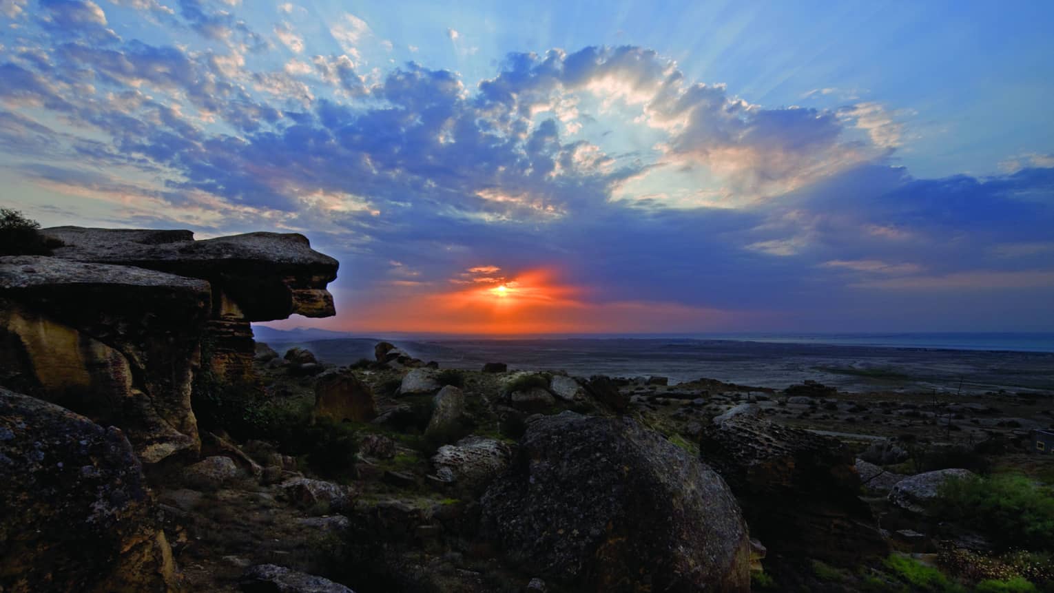 Sunbeams and fluffy azure blue clouds dance across a twilight sky, the vibrant red sun gently illuminating a rocky shoreline.