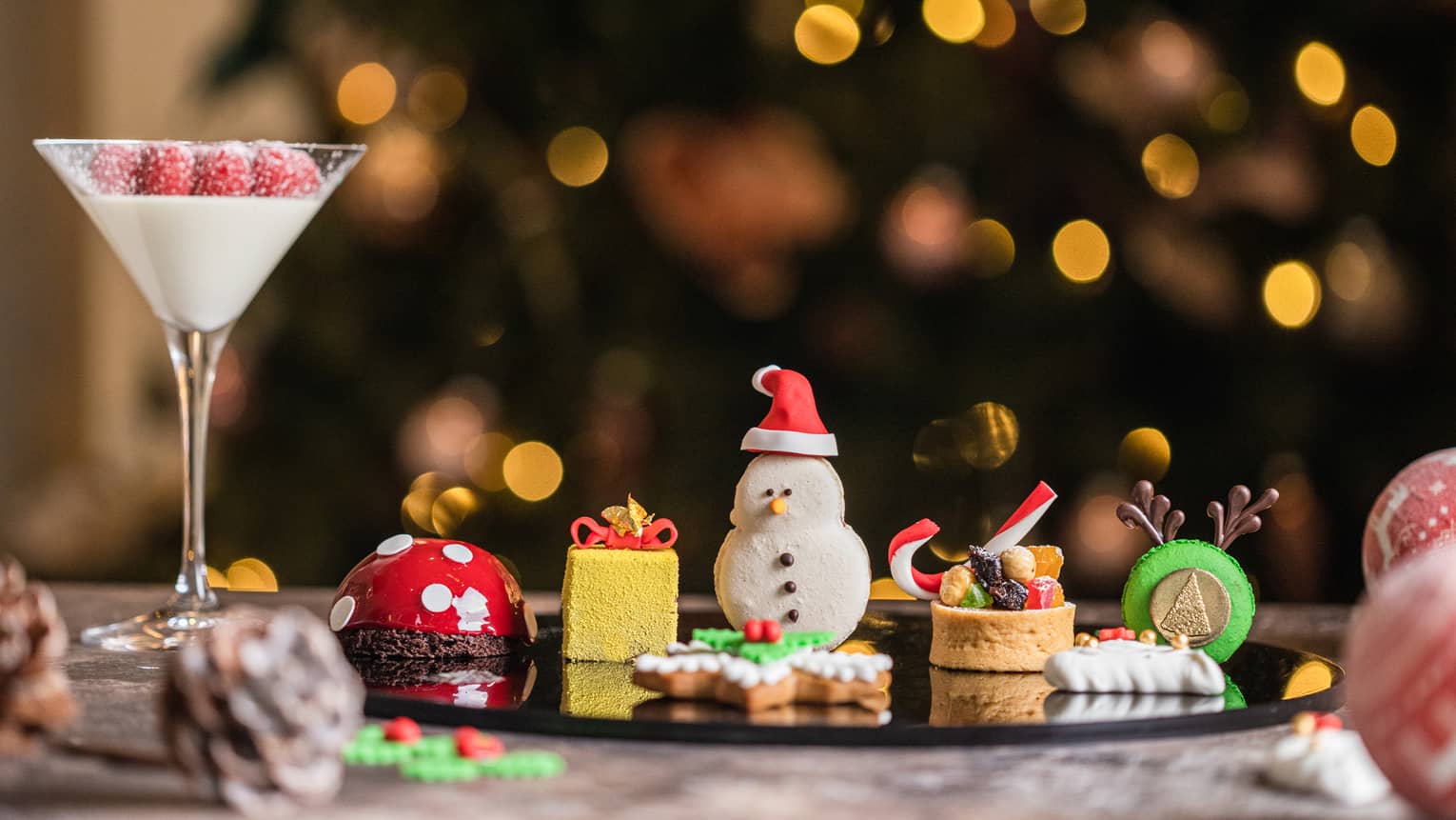 A row of holiday pstries including a cookie shaped like a snowman, a yellow cake shaped like a present box and a green cookie with reindeer antlers sits next to a martini glass filled with cream and berries