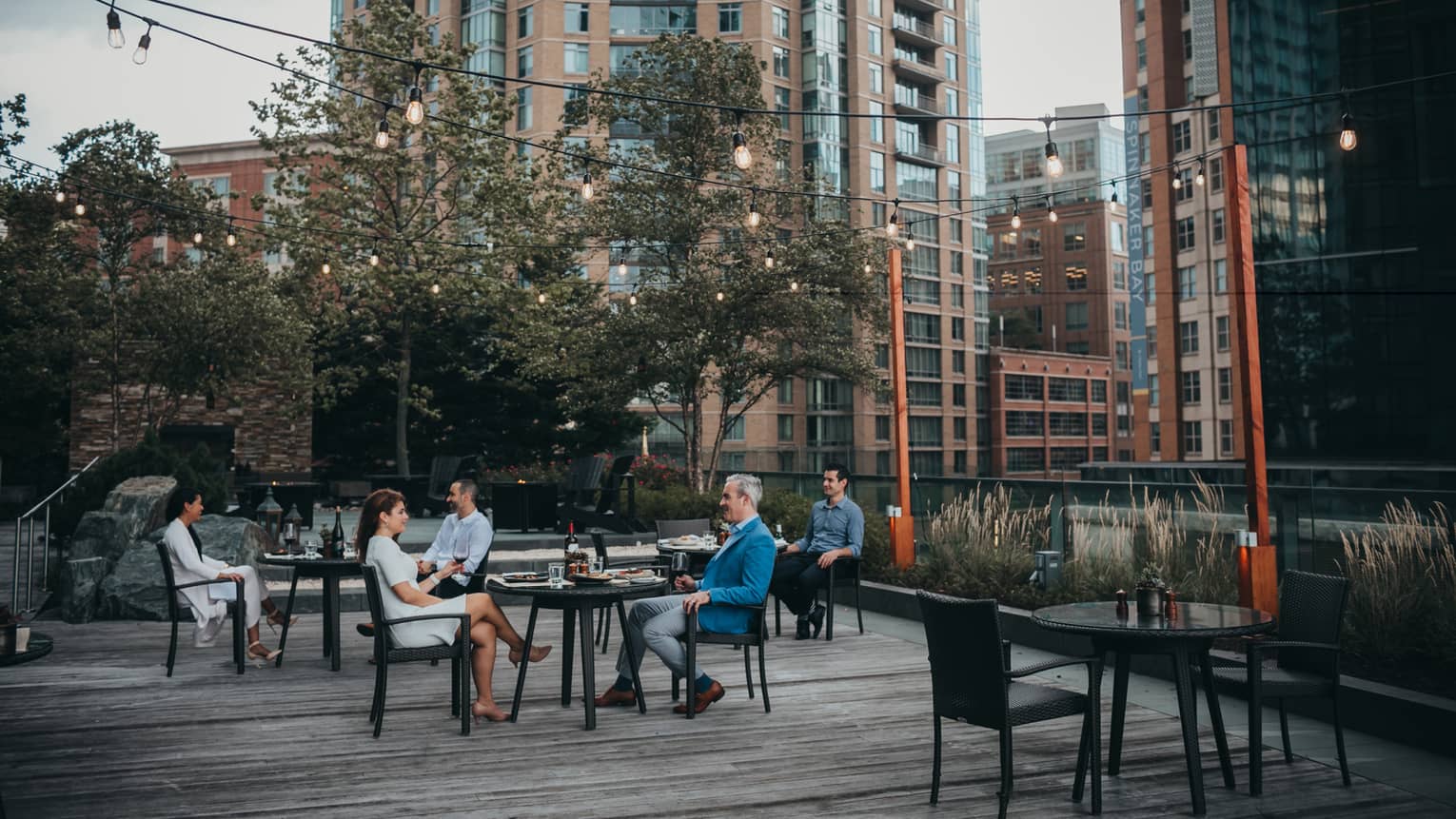 Three tables outside with a man and woman sitting at each enjoying a meal in the evening, there are trees and tall buildings in the background.