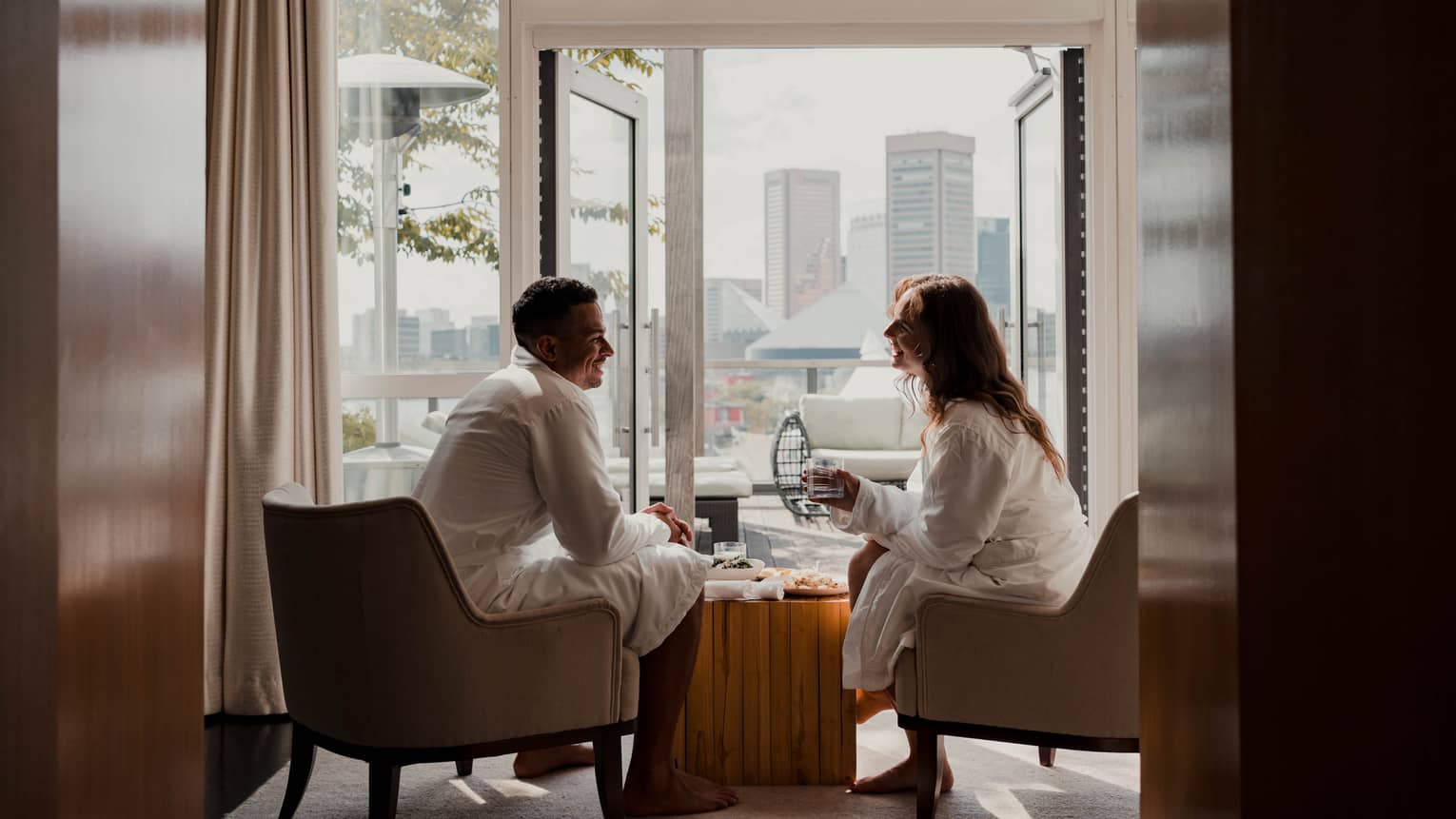 Two people in white robes sit in armchairs by an open balcony door with a cityscape view in the background, sharing a relaxed, casual conversation. 