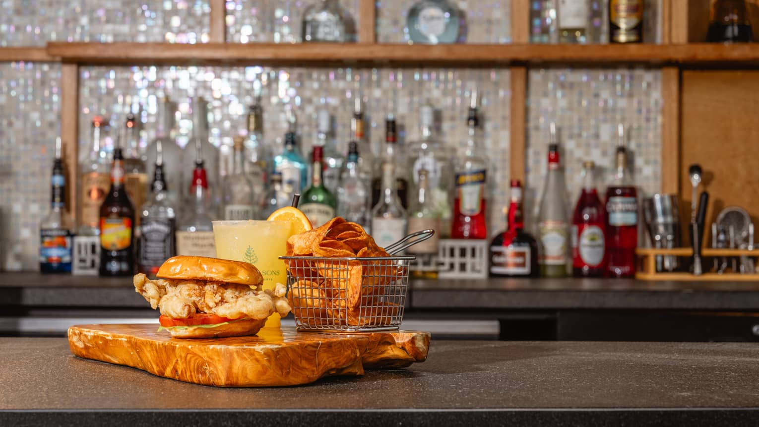 A wood serving tray with a sandwich, a side and a drink sits on a bar countertop.