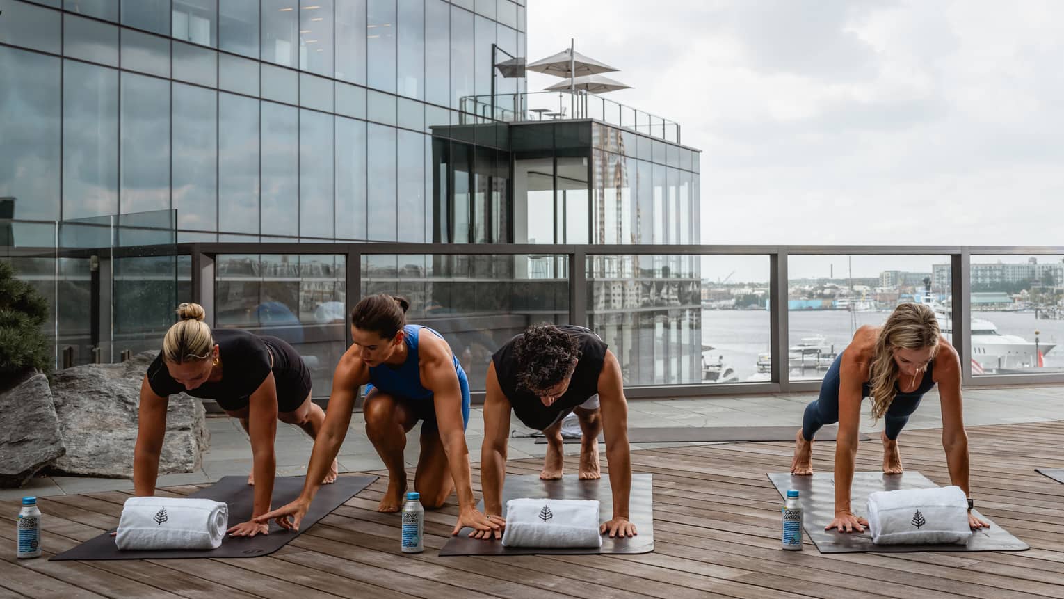 People in yoga poses on a rooftop deck, with city views in the background. An instructor gives guidance to two of the people.