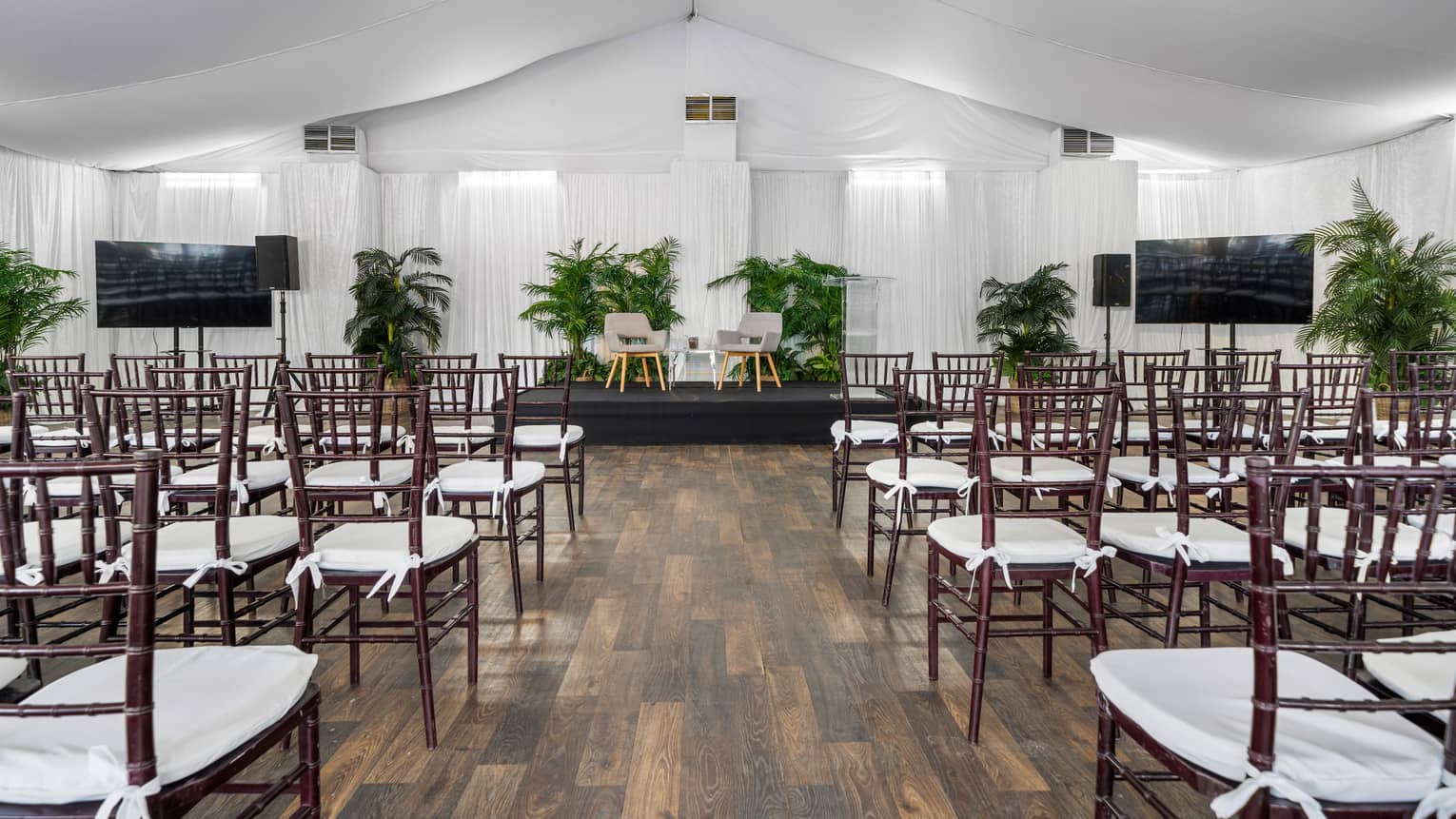 White event tent set up with brown cane chairs with white seat cushions in a theatre-style set up with a small black stage in front with two tan chairs and flat screen TVs on either side