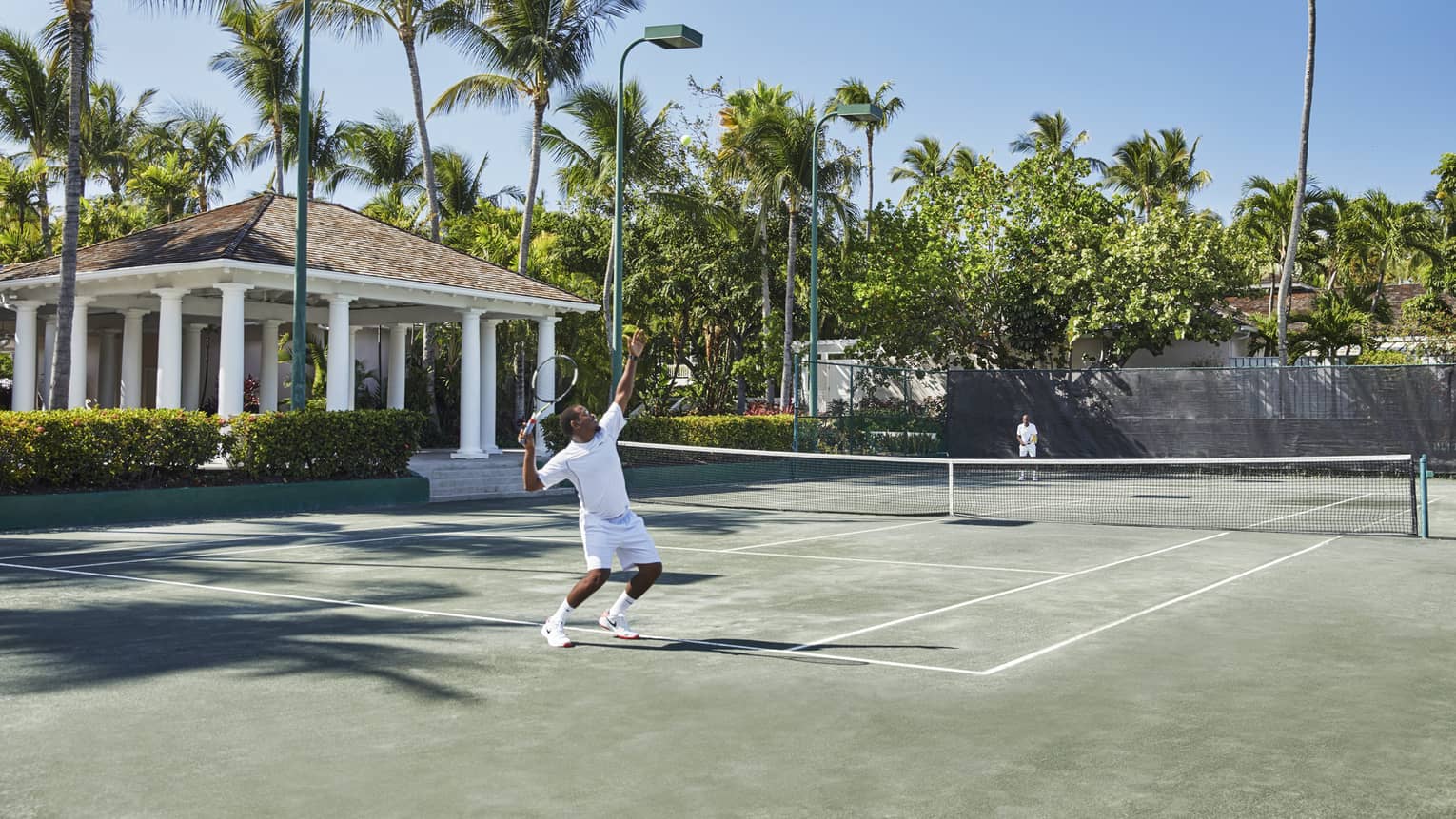 A man in all white trows a tennis ball high into the air, mid-serve 