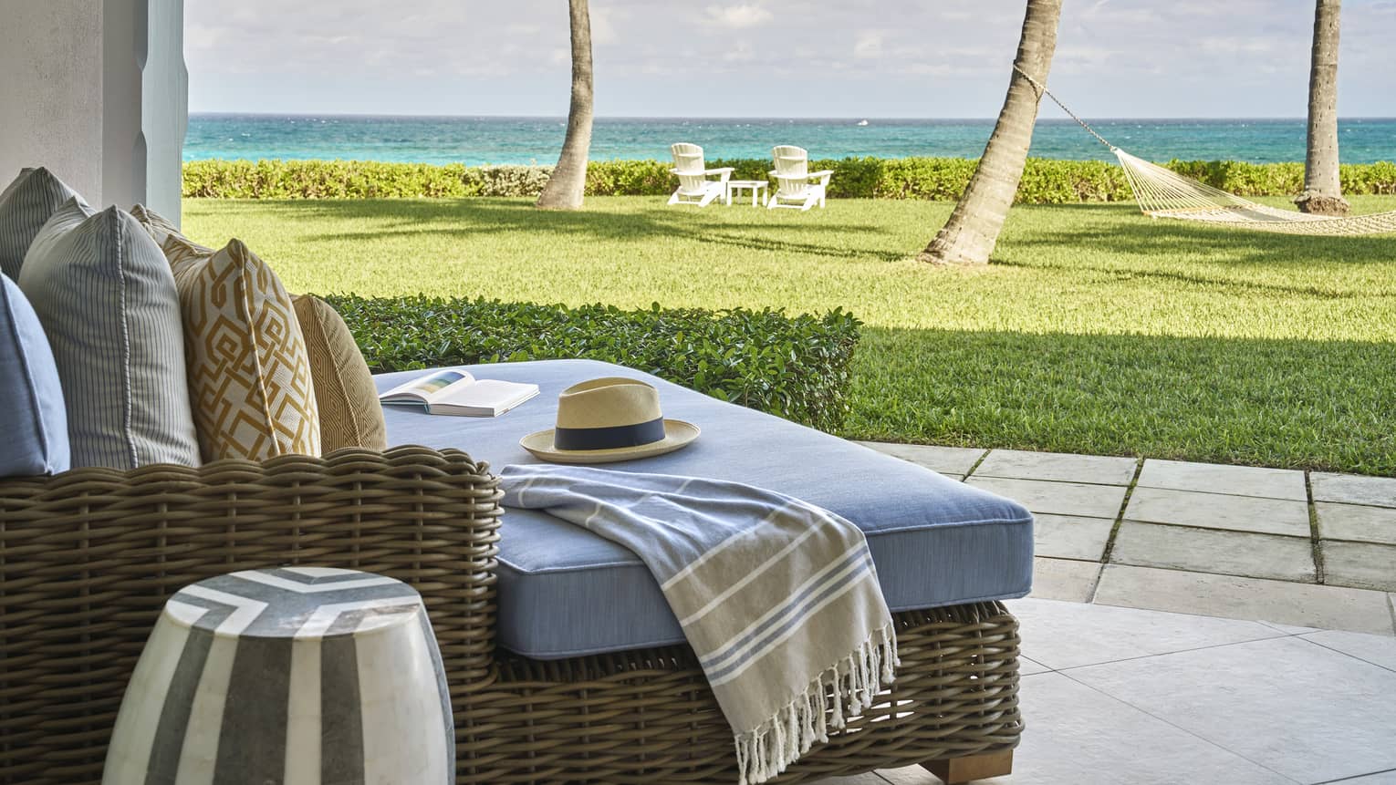 Resort room patio furnished with a blue upholstered daybed that looks out onto a green lawn with hammocks hanging between palm trees, two chairs and the ocean in the near distance