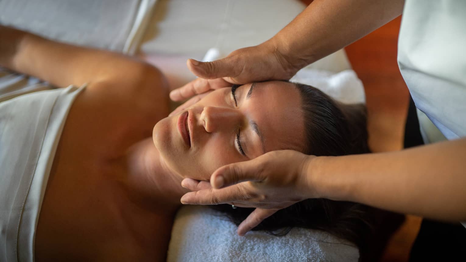 Woman lays on massage table with eyes closed as hands massage her cheeks and chin