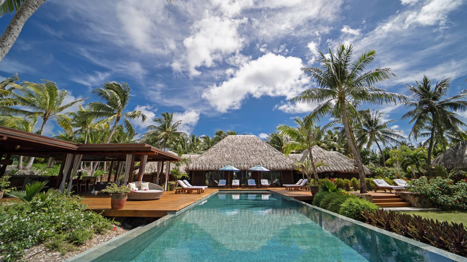 View from end of long rectangular pool, looking back across the pool to thatched-roof villa