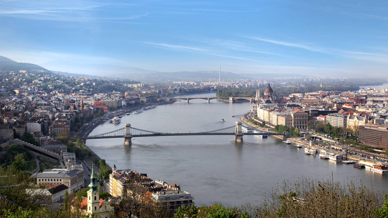 Aerial view of the Danube river skirted by two bridges, palatial buildings and densely built landscape receding uphill.