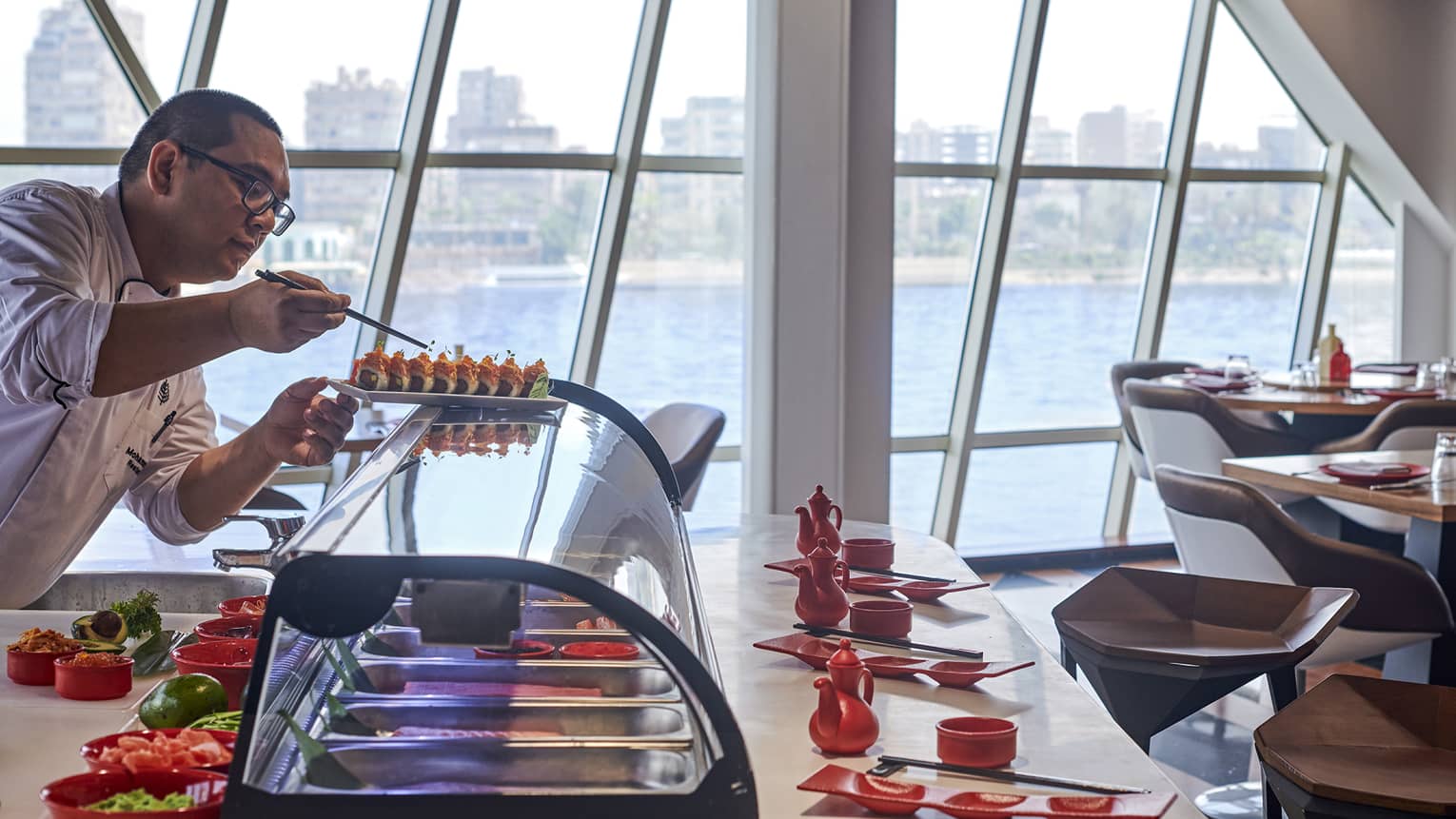 A chef carefully sets tiny greens onto crunchy roll sushi amid bowls, teapots and dipping trays, and panoramic harbour view.