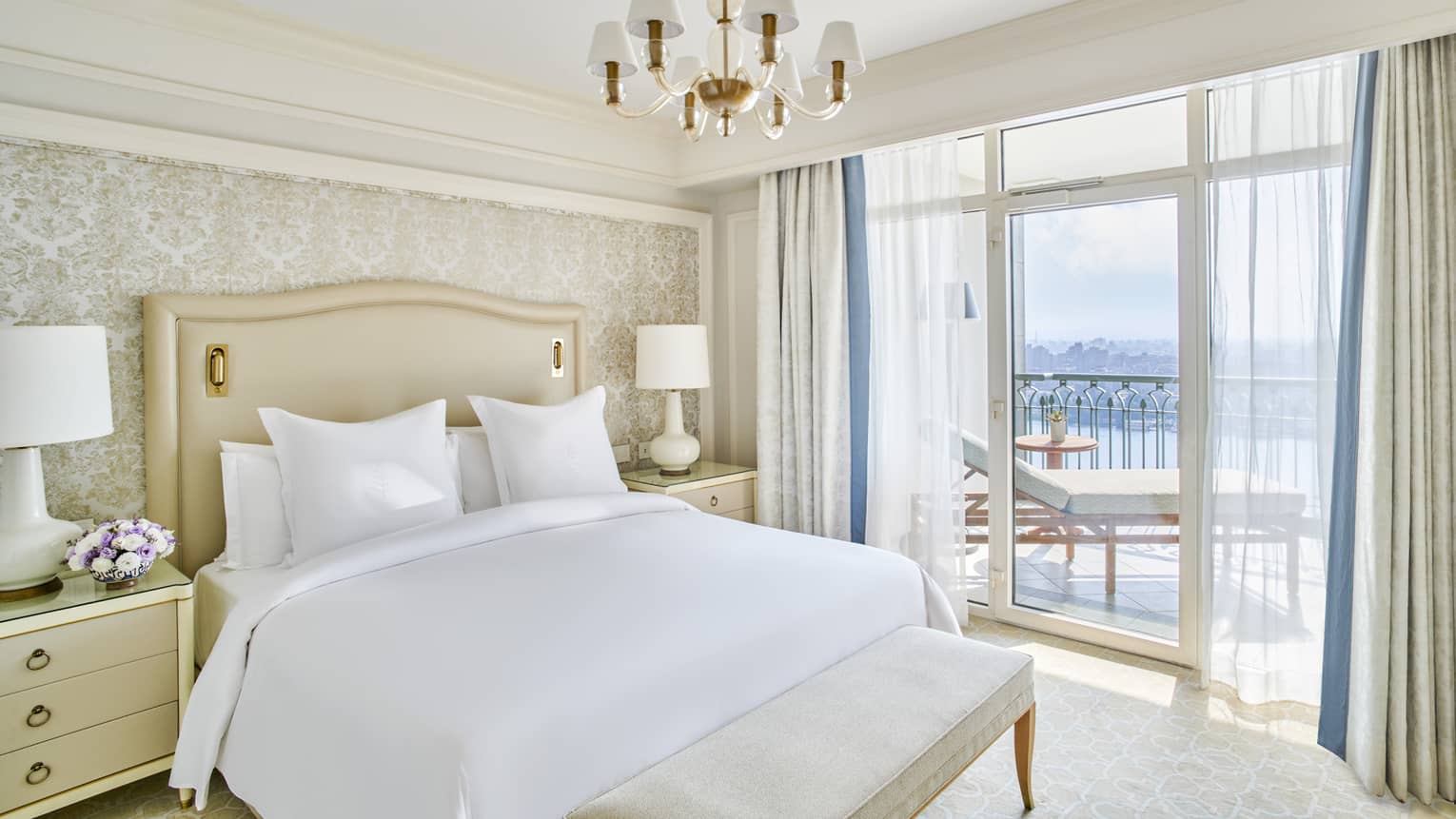 Guest bedroom with king bed, white linens and cream tufted headboard; foot bench, sliding glass door to balcony with Nile River views