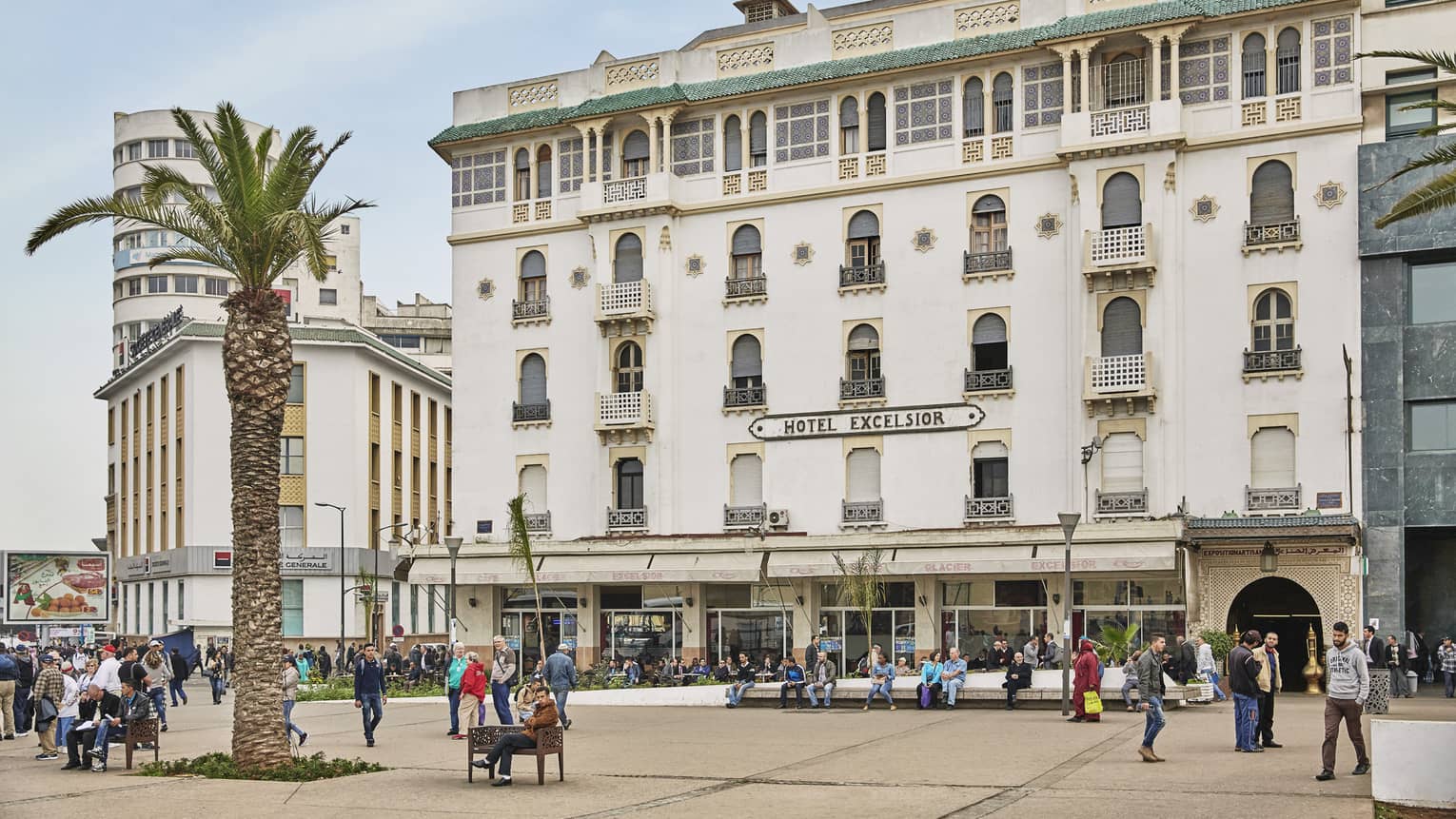People walk past palm tree along sidewalk under white building with sign Hotel Excelsior
