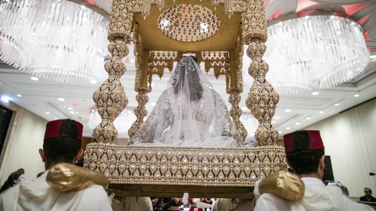 Wedding ceremony, back view of bride with white veil seated on beaded roofed platform carried by men