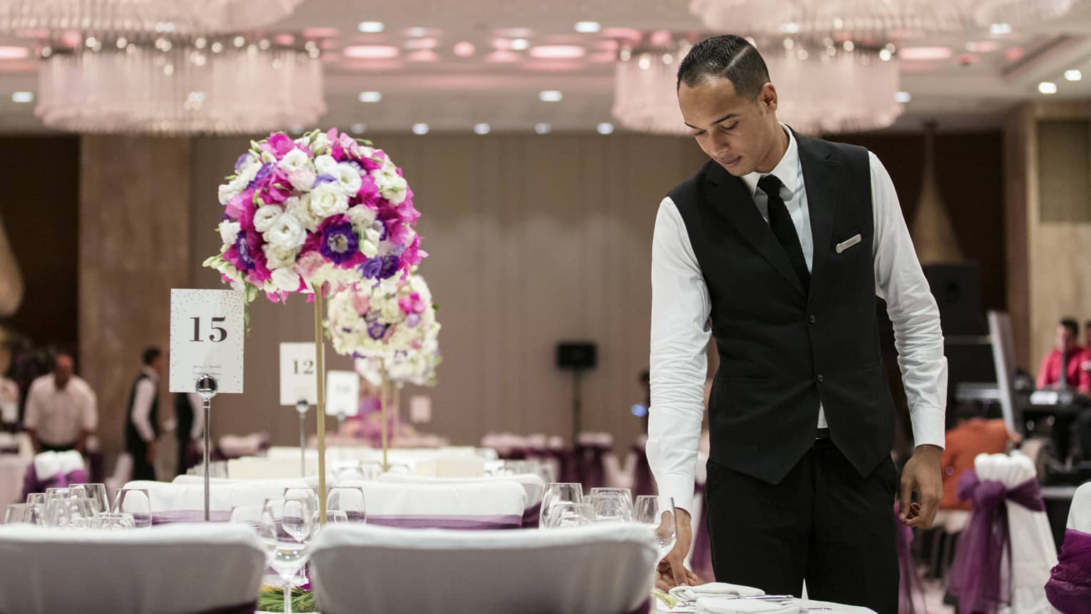 Hotel staff sets banquet table with white and purple floral arrangement, sign with number 15