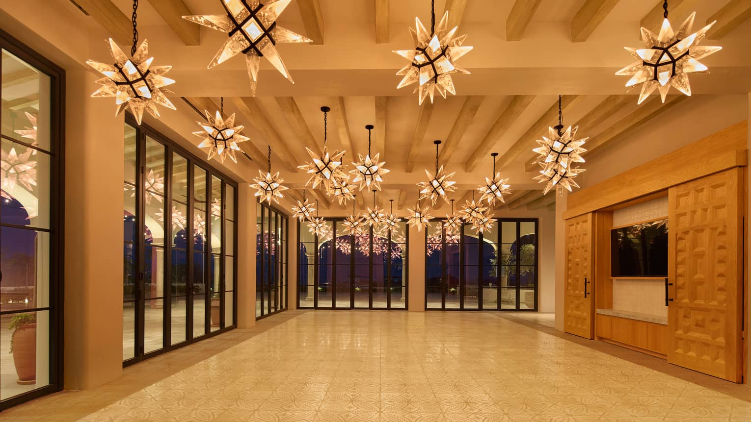Brightly lit and spacious indoor event space with warm neutral-colour interior, large flat-screen TV with decorative sliding panels, floor-to-ceiling windows, and star-shaped pendant lighting
