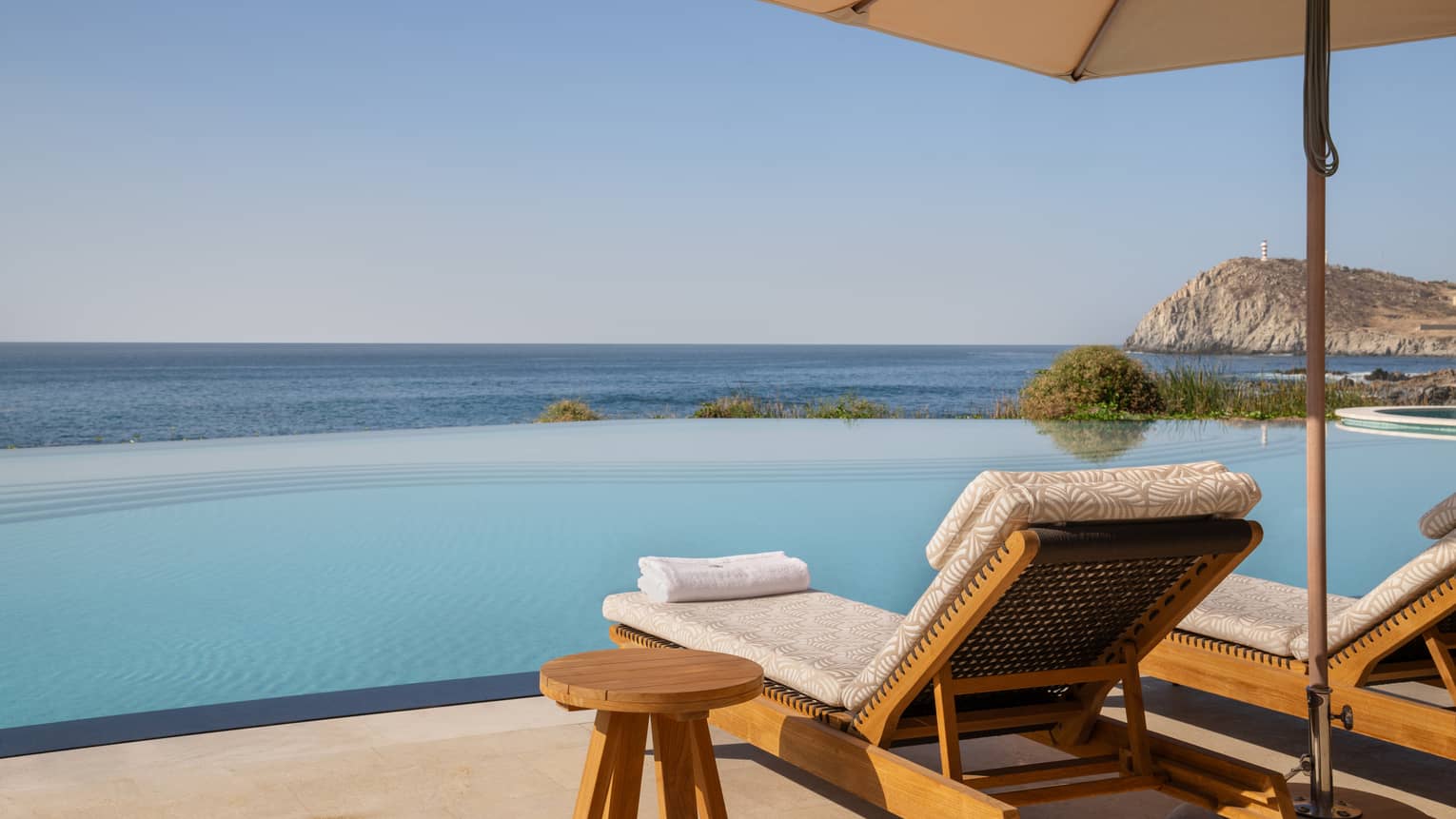 Two lounge chairs placed beneath a cream umbrella sit next to an infinity pool that overlooks the ocean