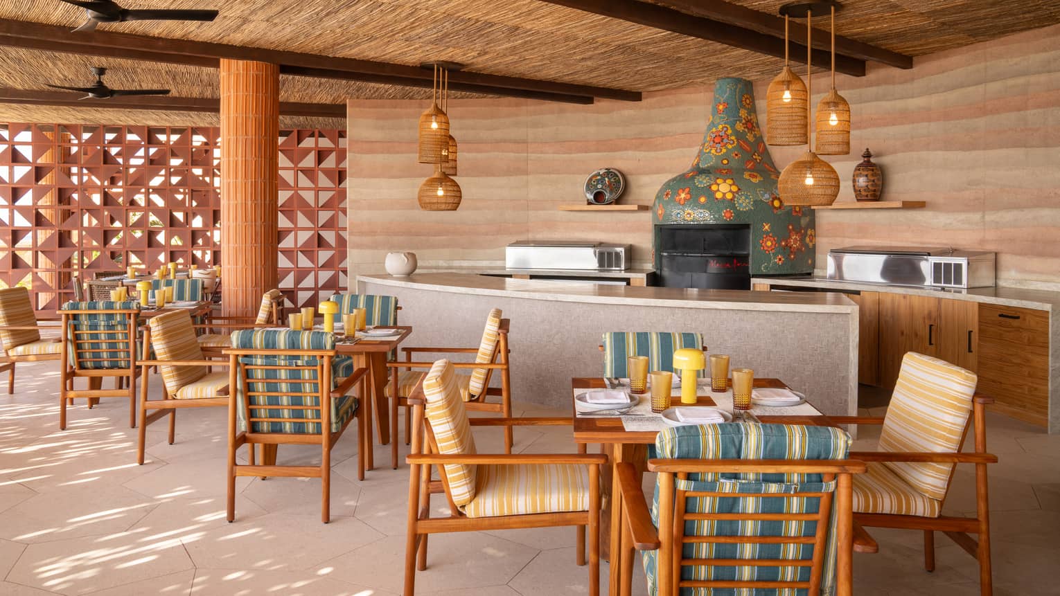 Outdoor kitchen with colourful tile oven, thatched ceilings above, light-grey counters and three dining tables in front