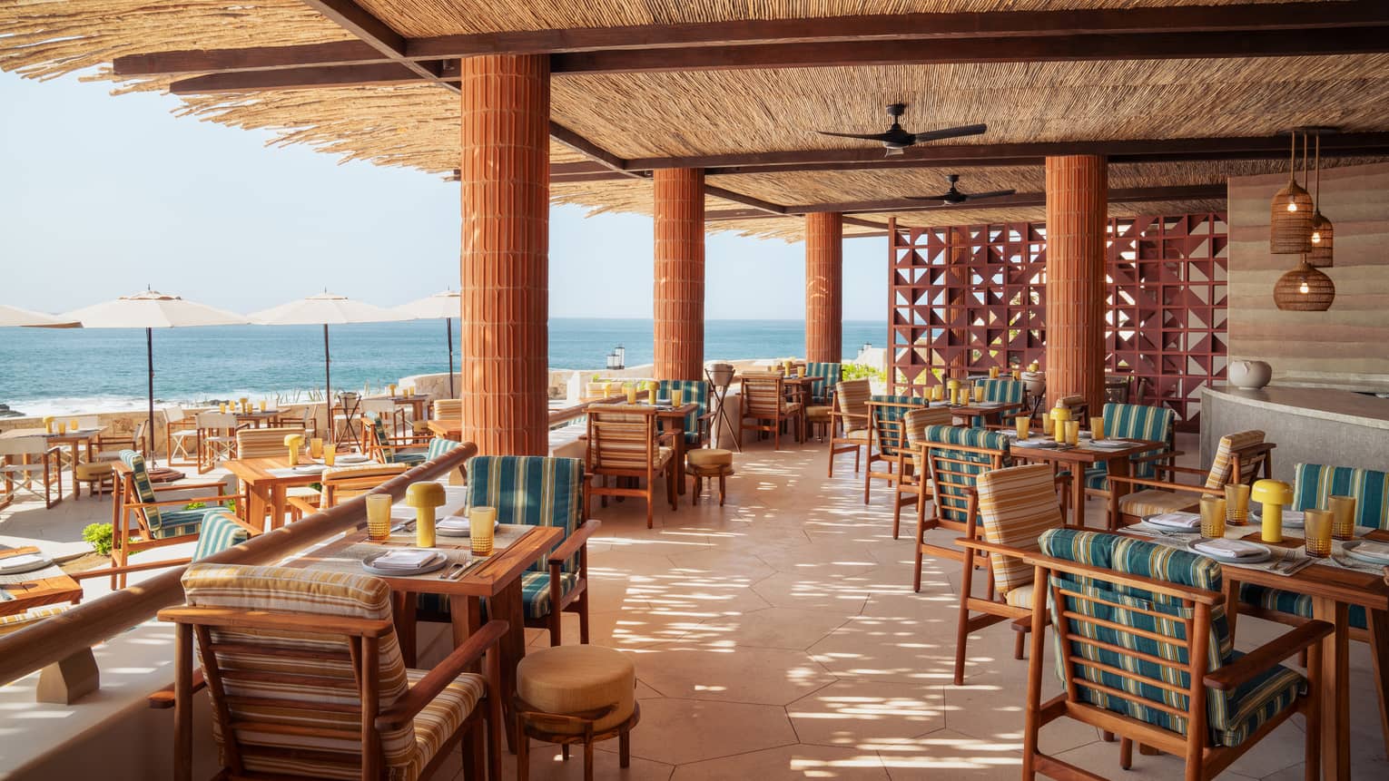Covered outdoor dining terrace with two rows of table beneath a thatched roof with large wood pillars and dark-wood beams, with the ocean in the background