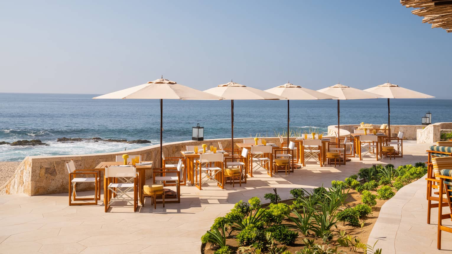 Open-air terrace with a row of tables and umbrellas set along a curved stone wall overlooking the ocean