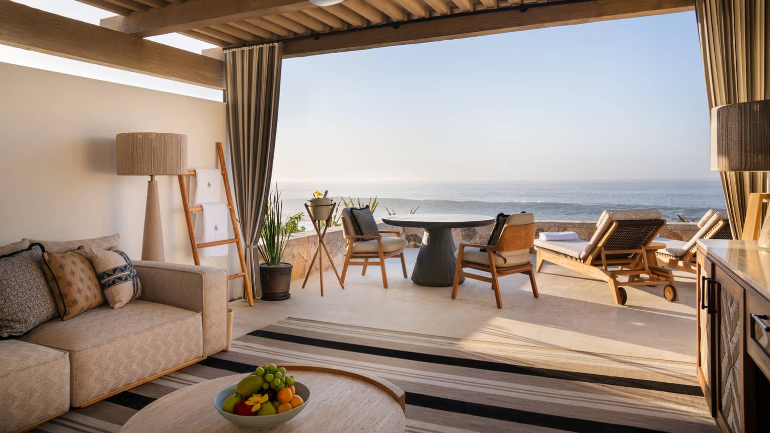 Oceanfront cabana with beige sofa, lamp and round wood coffee table under a roof with a round table and chairs and two lounge chairs out on the open-air patio overlooking the ocean