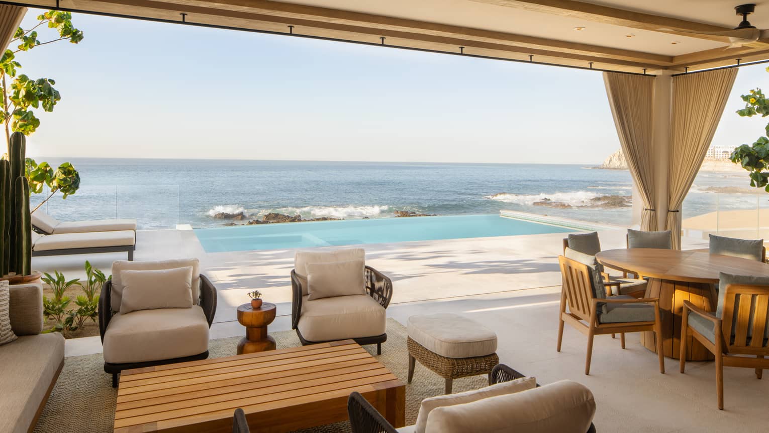 Luxury villa terrace with seating, a dining table and an infinity pool overlooking the ocean, surrounded by greenery and lounge chairs