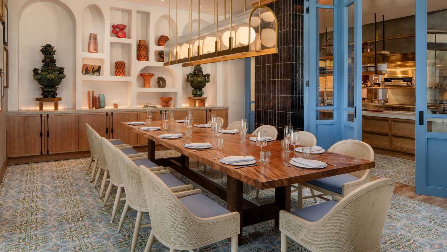 Private dining room with a wood, rectangular table, 11 cream-coloured chairs and a rectangular light fixture overhead, with a wall of arched shelving filled with statues as the backdrop and bright blue doors on the side