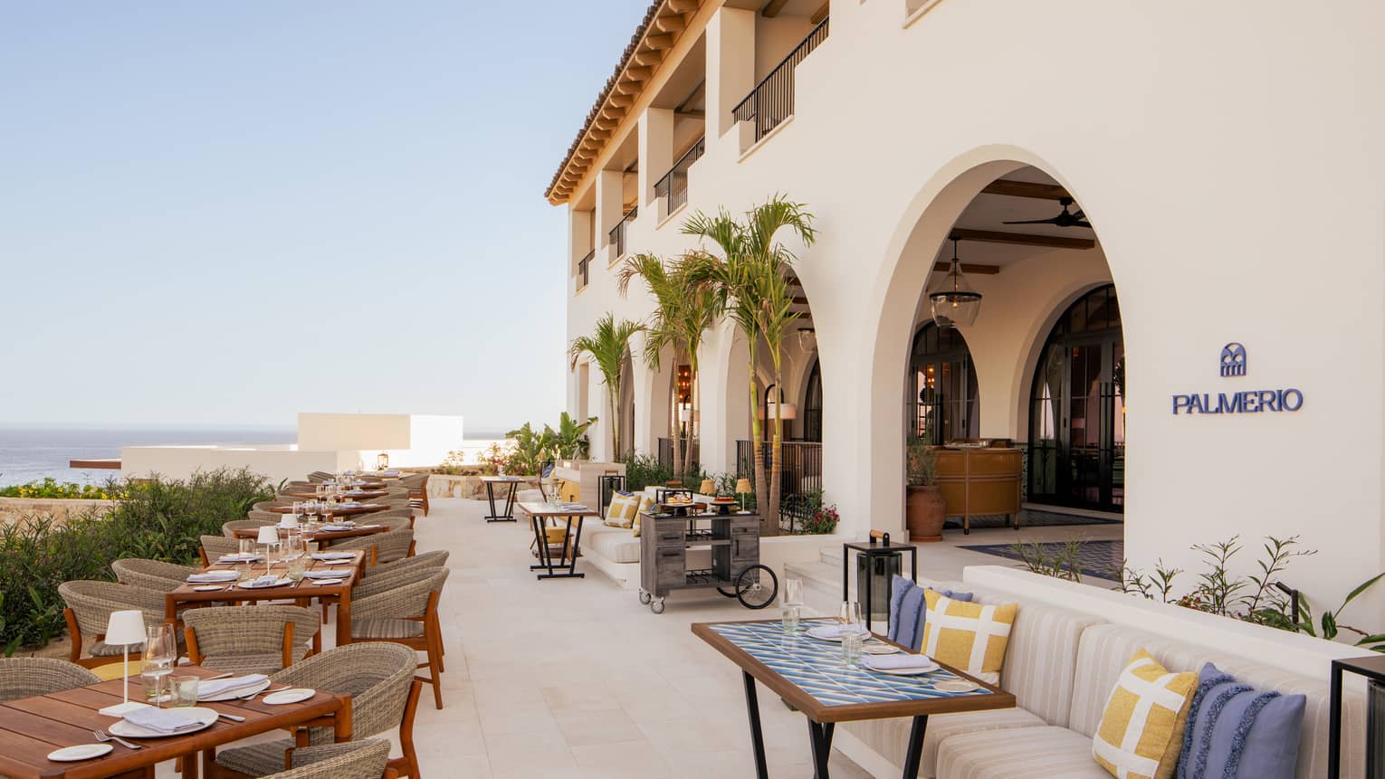 Open-air restaurant terrace with tables lining the edge and booth seating on the other with the ocean in the distance