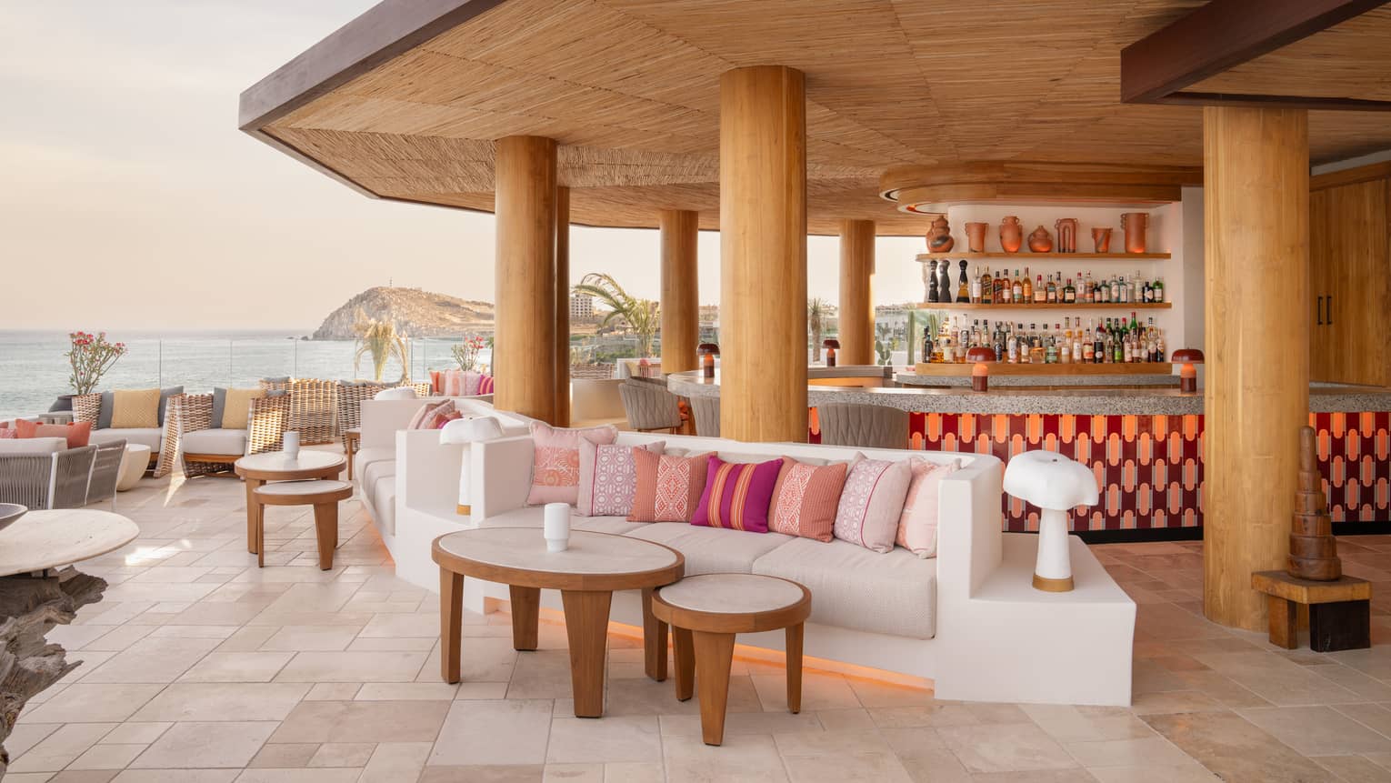 Rooftop bar with large round counter, wooden pillars, white and pink lounge seating and views overlooking the ocean