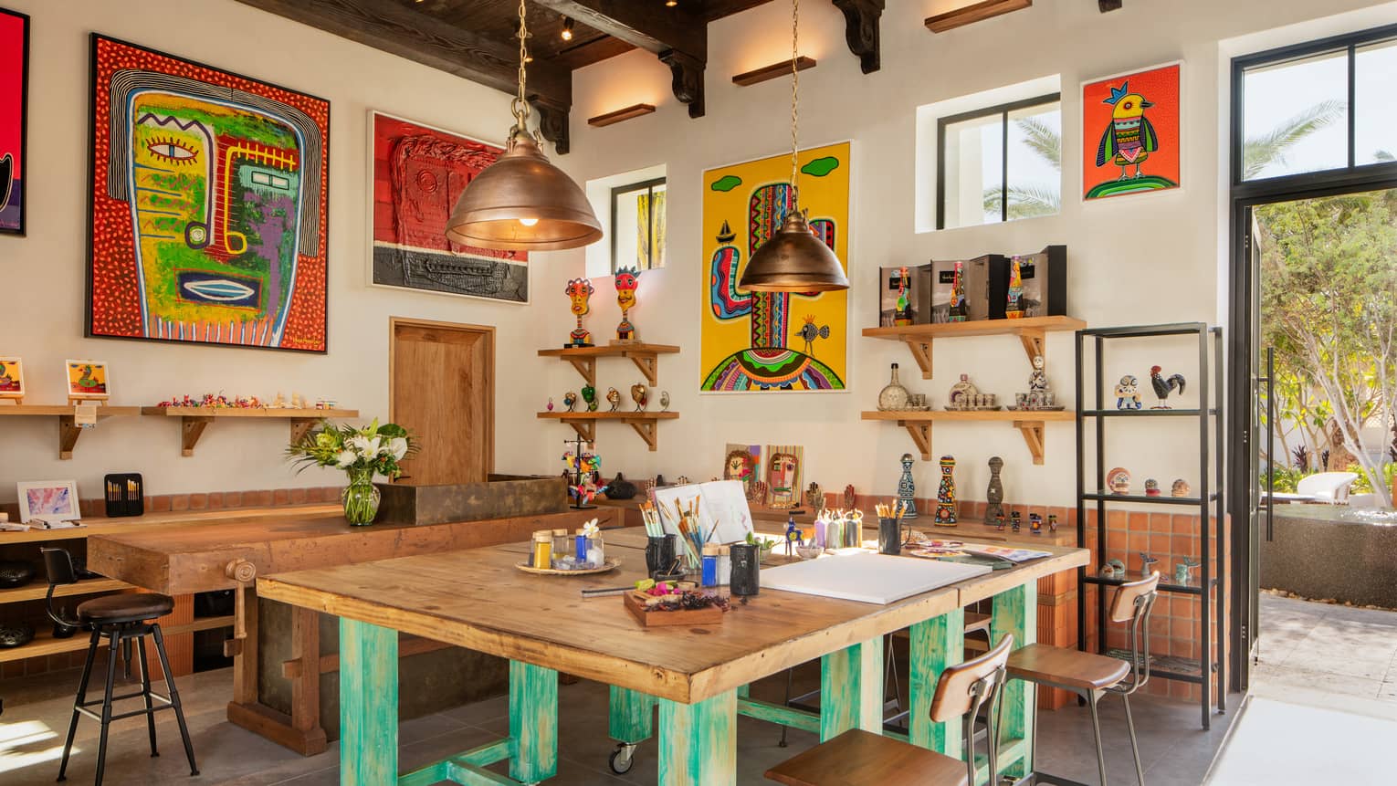 A bright, airy art studio with a large square wooden worktable, shelves of knick-knacks and colourful paintings on the walls.