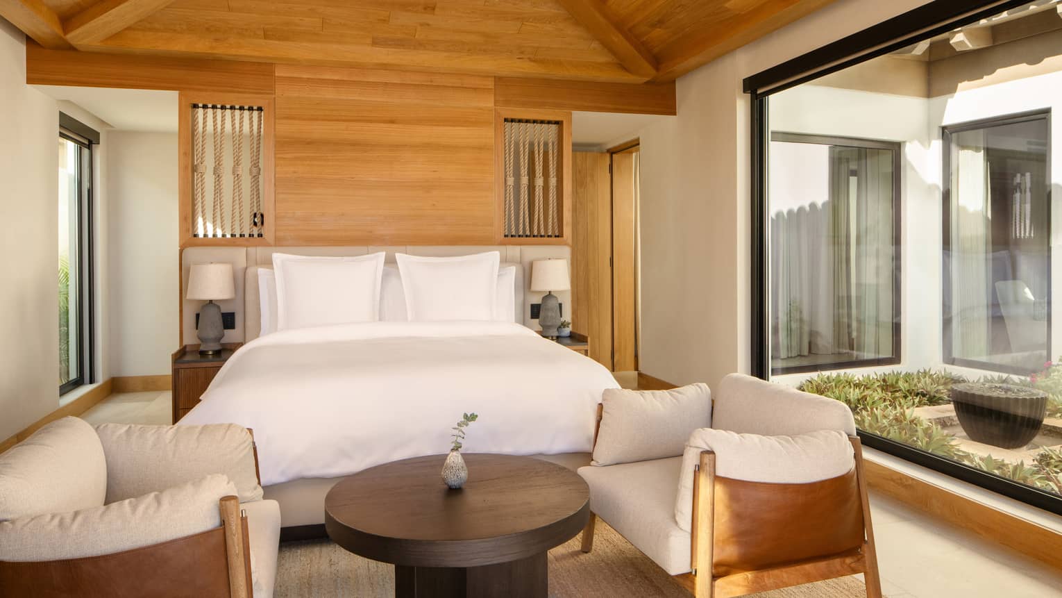 Bedroom with king bed, wooden vaulted ceiling, two arm chairs facing a round coffee table, and a wall of window