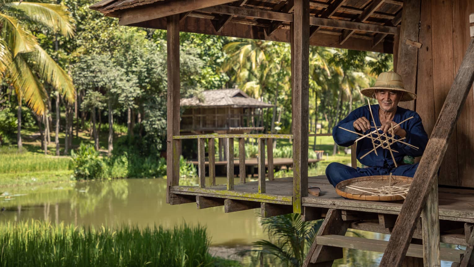 Man weaves a traditional Thai basket on the porch of a riverside pagoda
