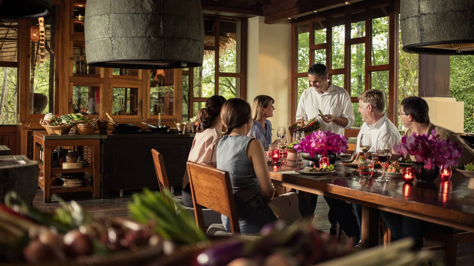 Group dining indoors with a chef presenting ingredients, surrounded by flowers, candles and tropical wooden decor.