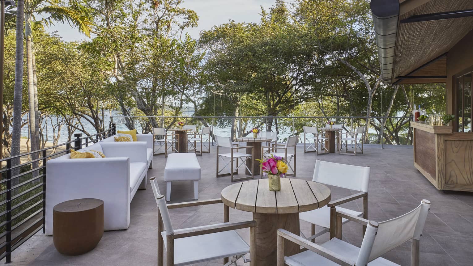 Outdoor terrace overlooking the beach with white sofas, various wooden tables and white chairs and an outdoor wooden bar