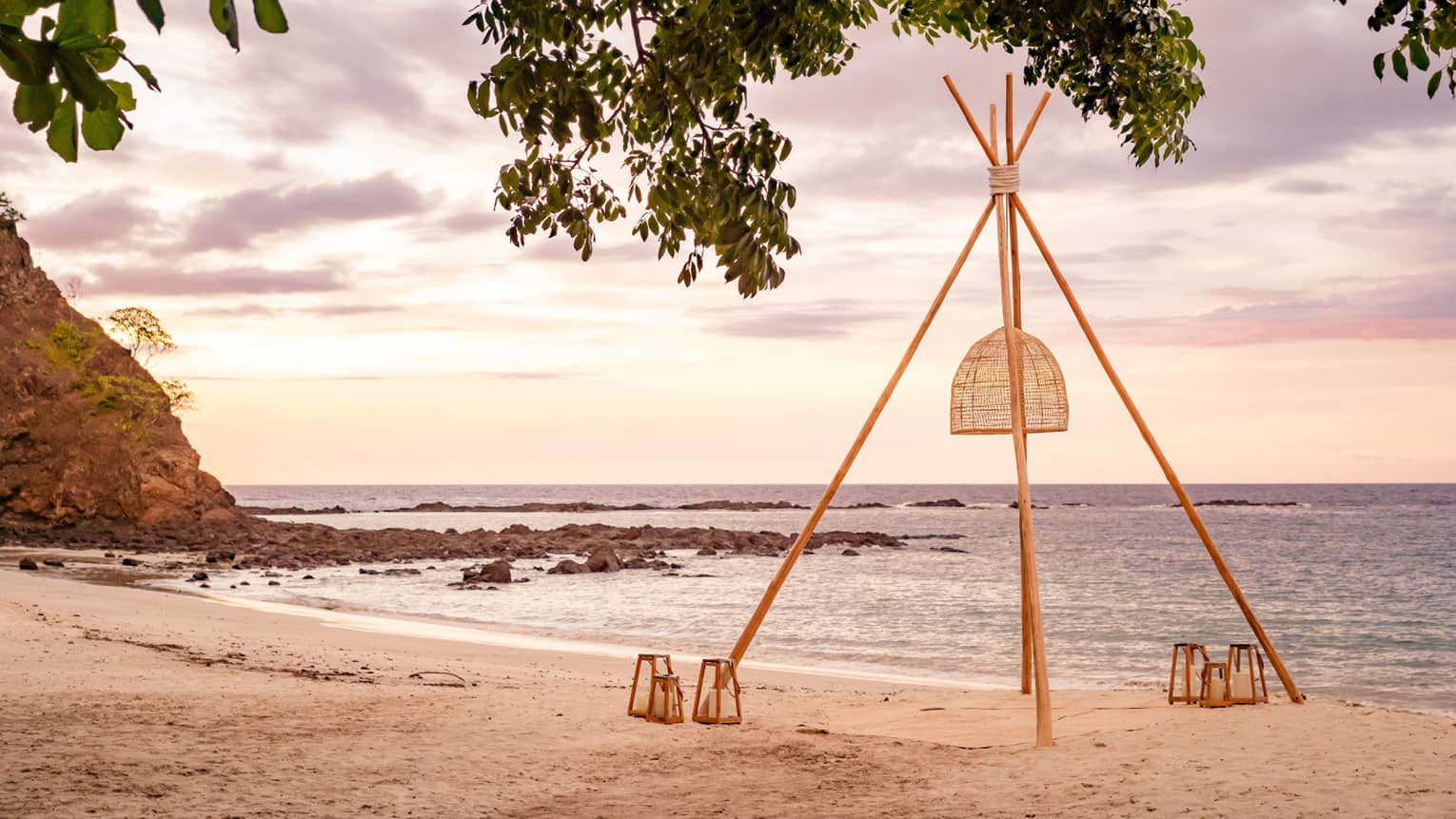 Four poles set up to hold a rattan lantern with other lanterns set on the ground on a private beach with the ocean nearby
