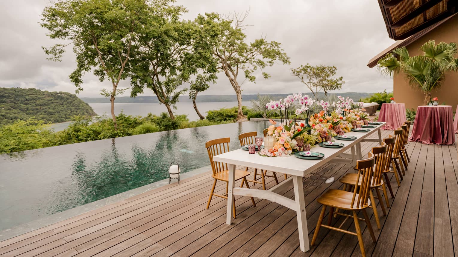 White rectangular dining table set for 12 with a large floral centrepiece on a wooden deck next to a private infinity-edge pool with trees and the ocean seen in the distance